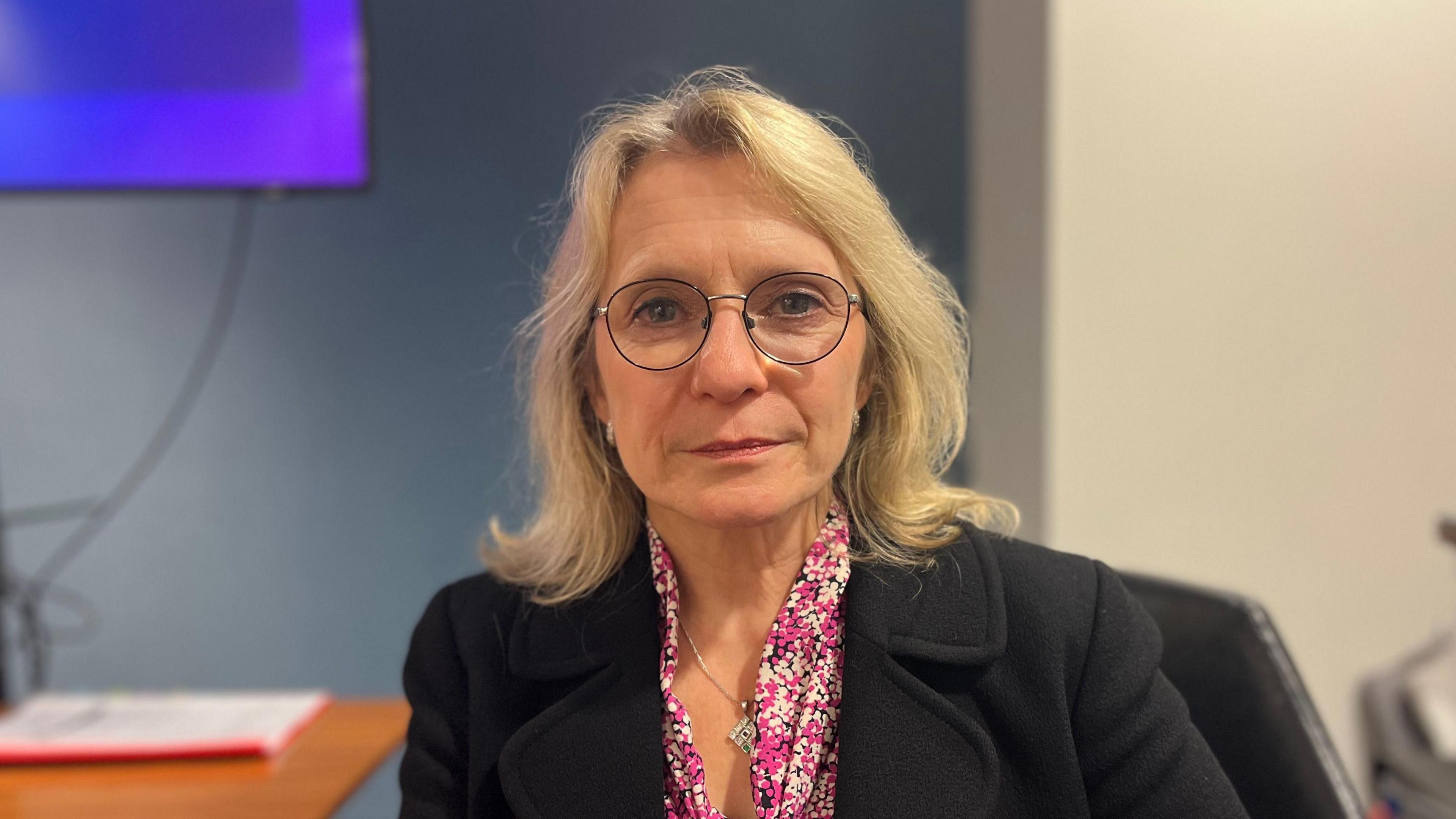 Elaine Millar is looking at the camera. She is a middle-aged white woman with blonde hair to her shoulders. She is wearing black wire-rimmed glasses, a pink and white blouse and a black jacket. The background is an out of focus room behind her.