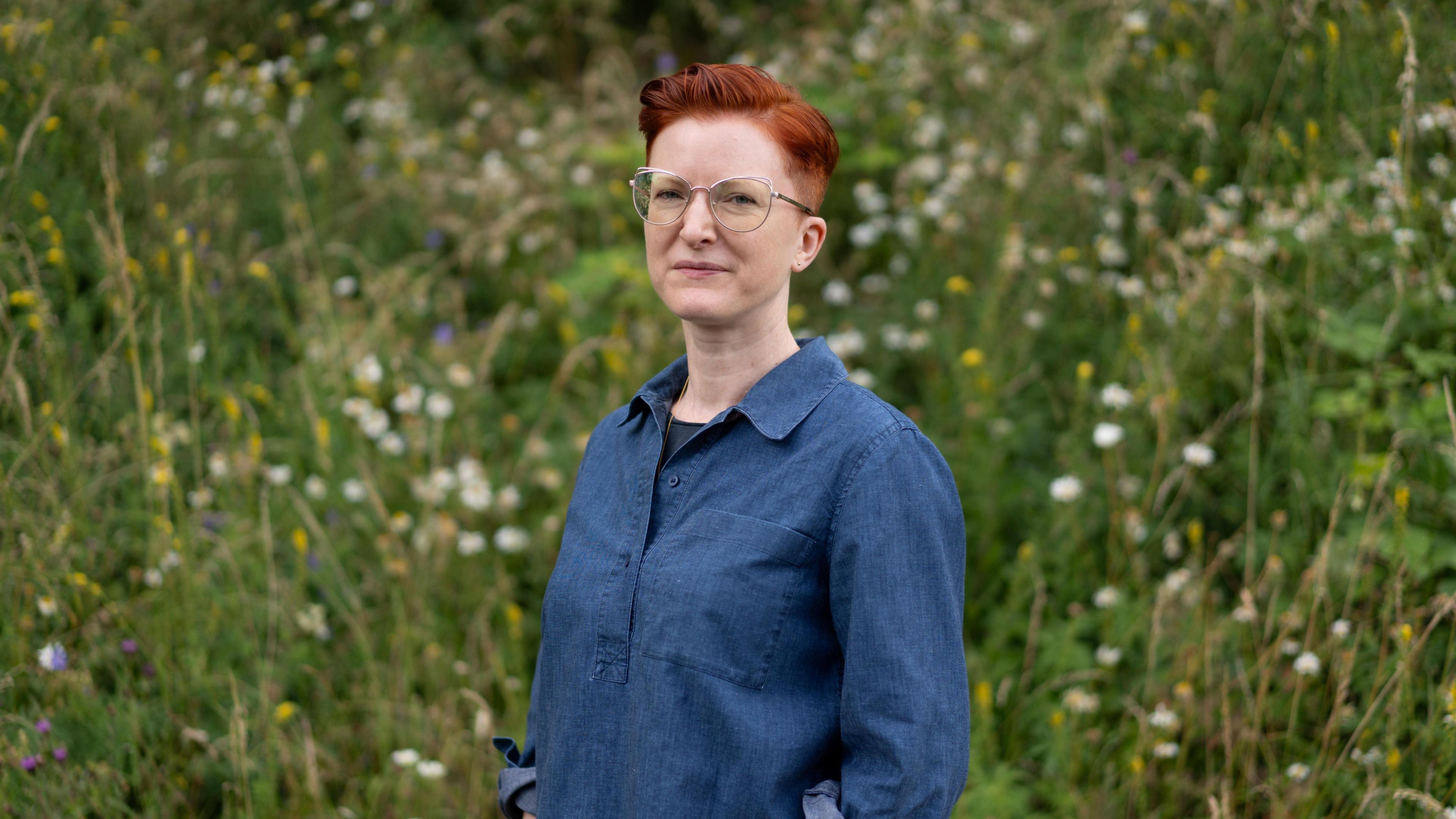 Rosie is standing in front of a meadow of wildflowers and wearing a denim shirt, with short red hair and glasses, looking at the camera with a neutral expression.