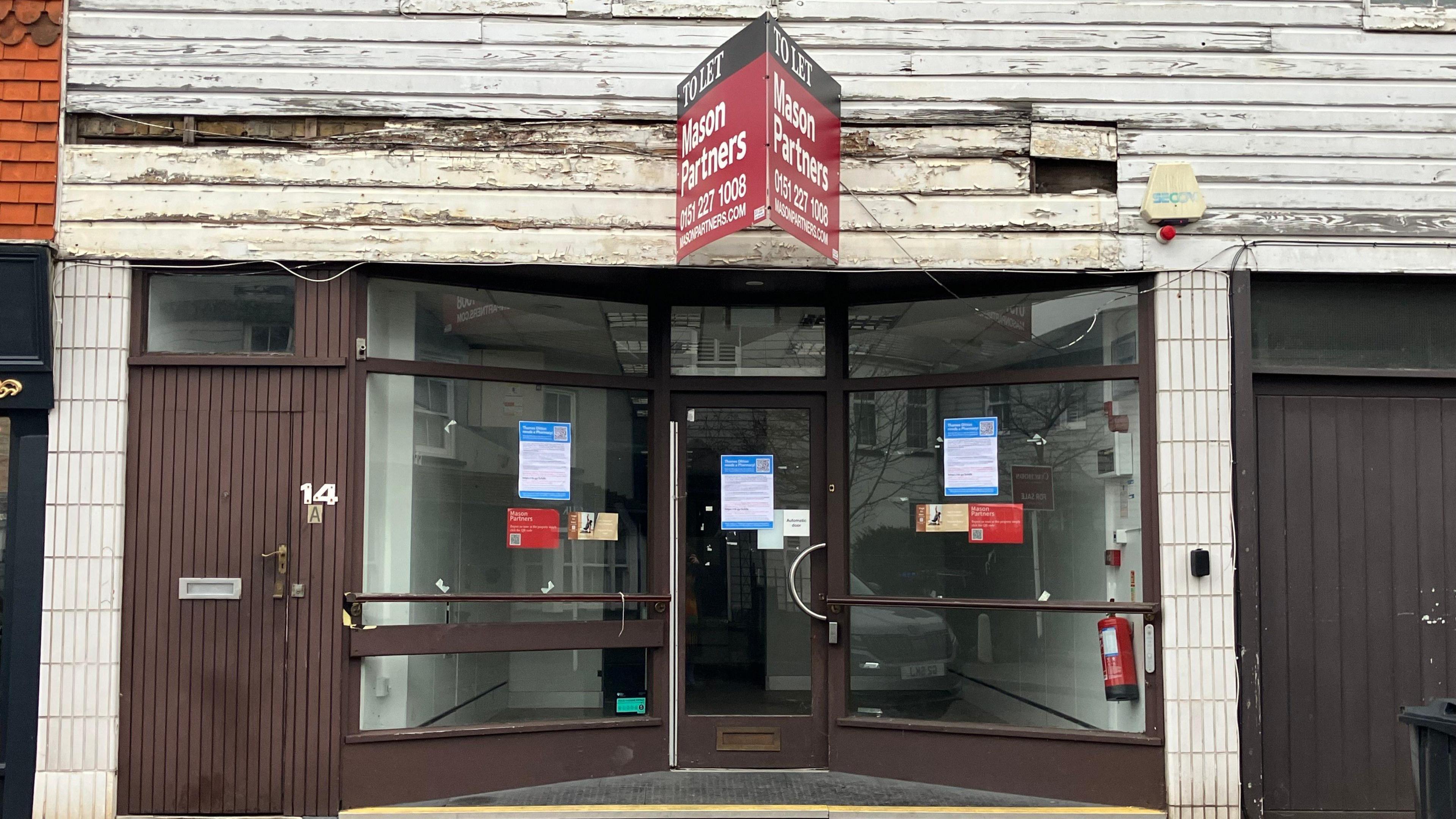 The image shows a front building view of a former Boots pharmacy in Thames Ditton. The door and window frames are painted brown, with the number 14 next to the door frame, with the letter "A" underneath it. There are old slabs of wood painted white above the doorframe, with some of them completely gone.