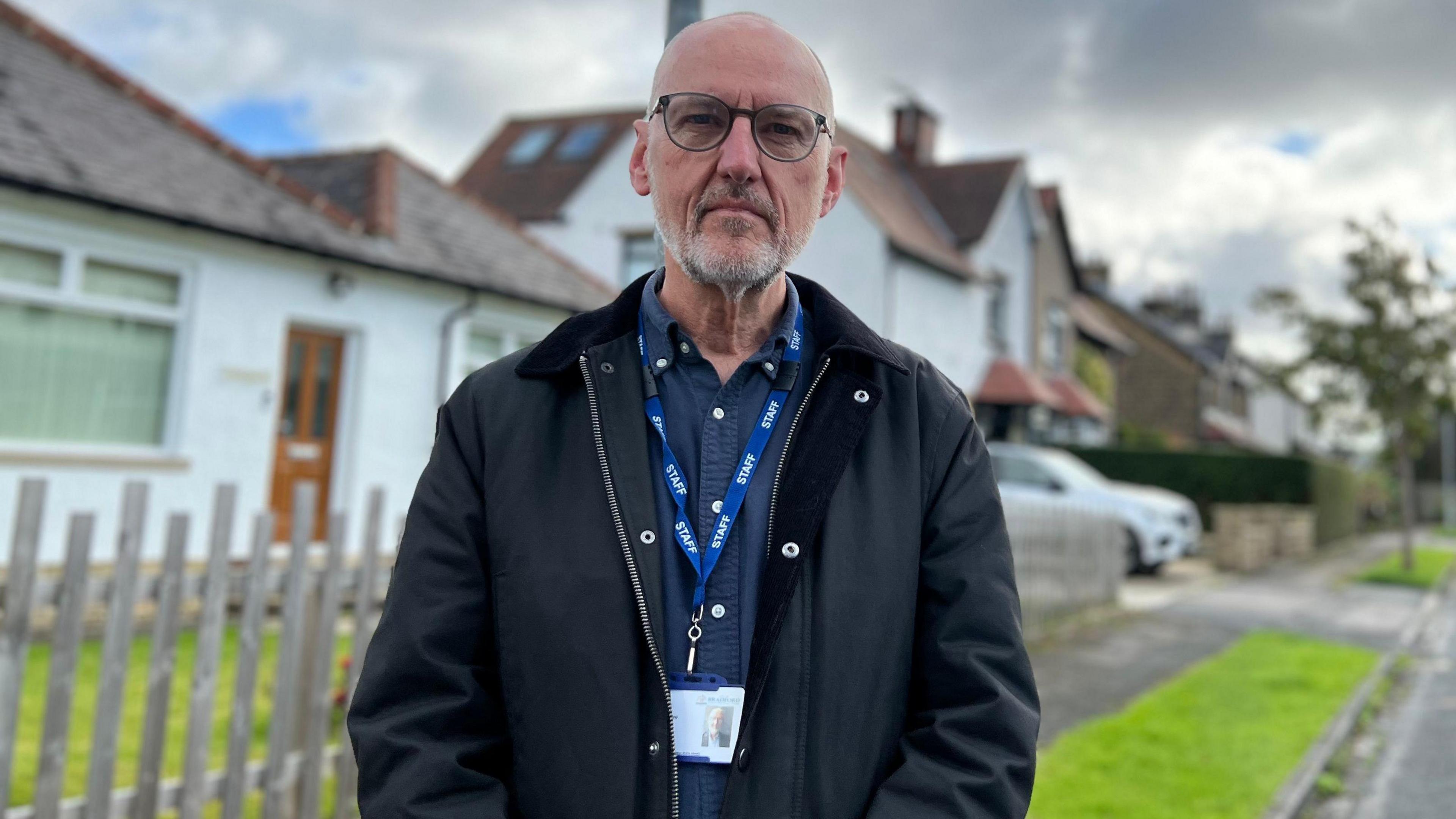 A man stands in a pretty looking suburban street with white painted houses and neatly trimmed grass verges visible  in the background. He is middle aged, has a shaven head, a short, stubbly white beard and black glasses. He wears a dark blue shirt, and dark zip up jacket with collar.