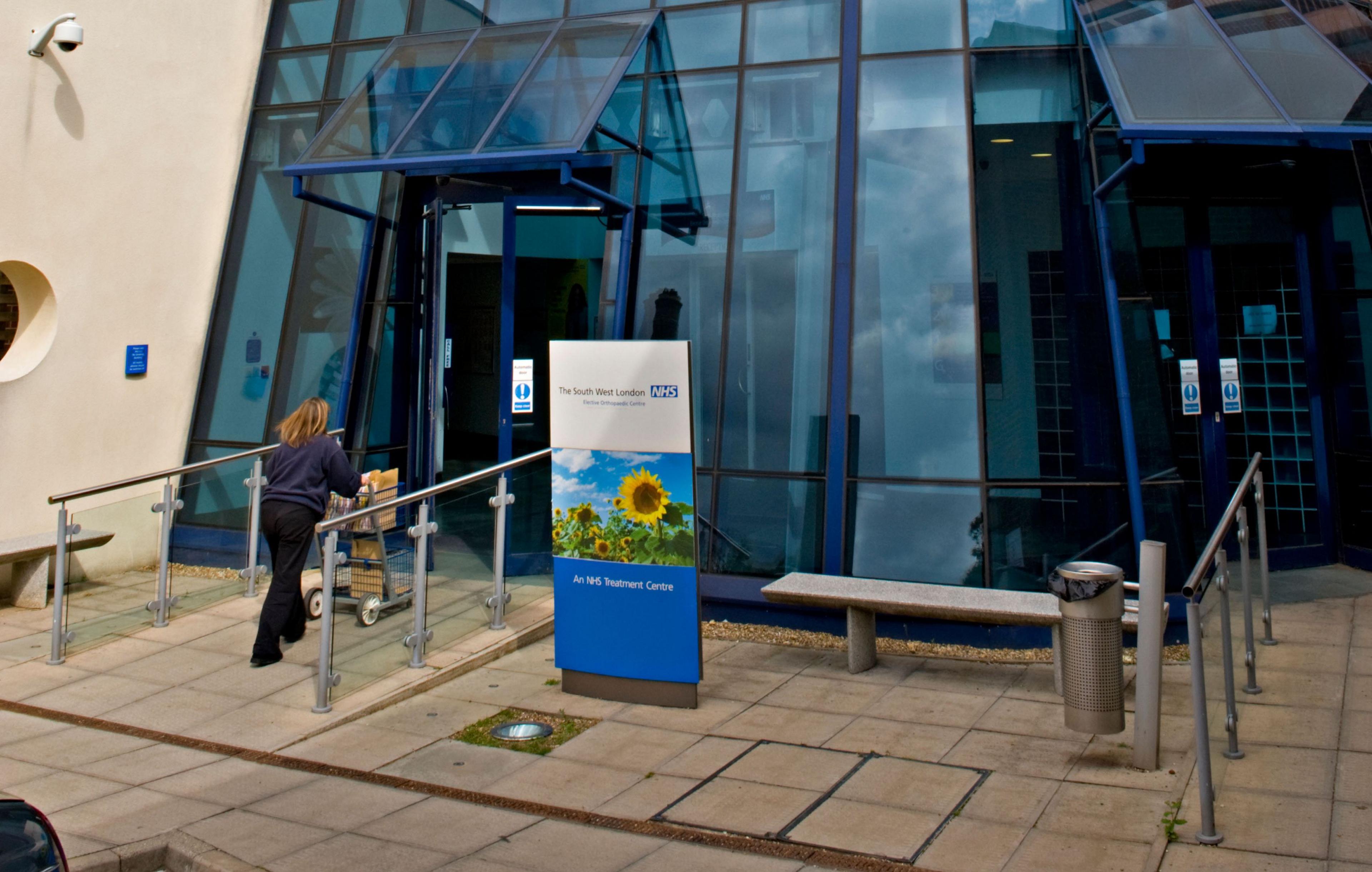 An outdoor shot of the entrance of the South West London Elective Orthopaedic Centre, a glass building with an NHS sign at the front, with nn employee walking up to the door pushing a trolley, taken in Epsom in 2009