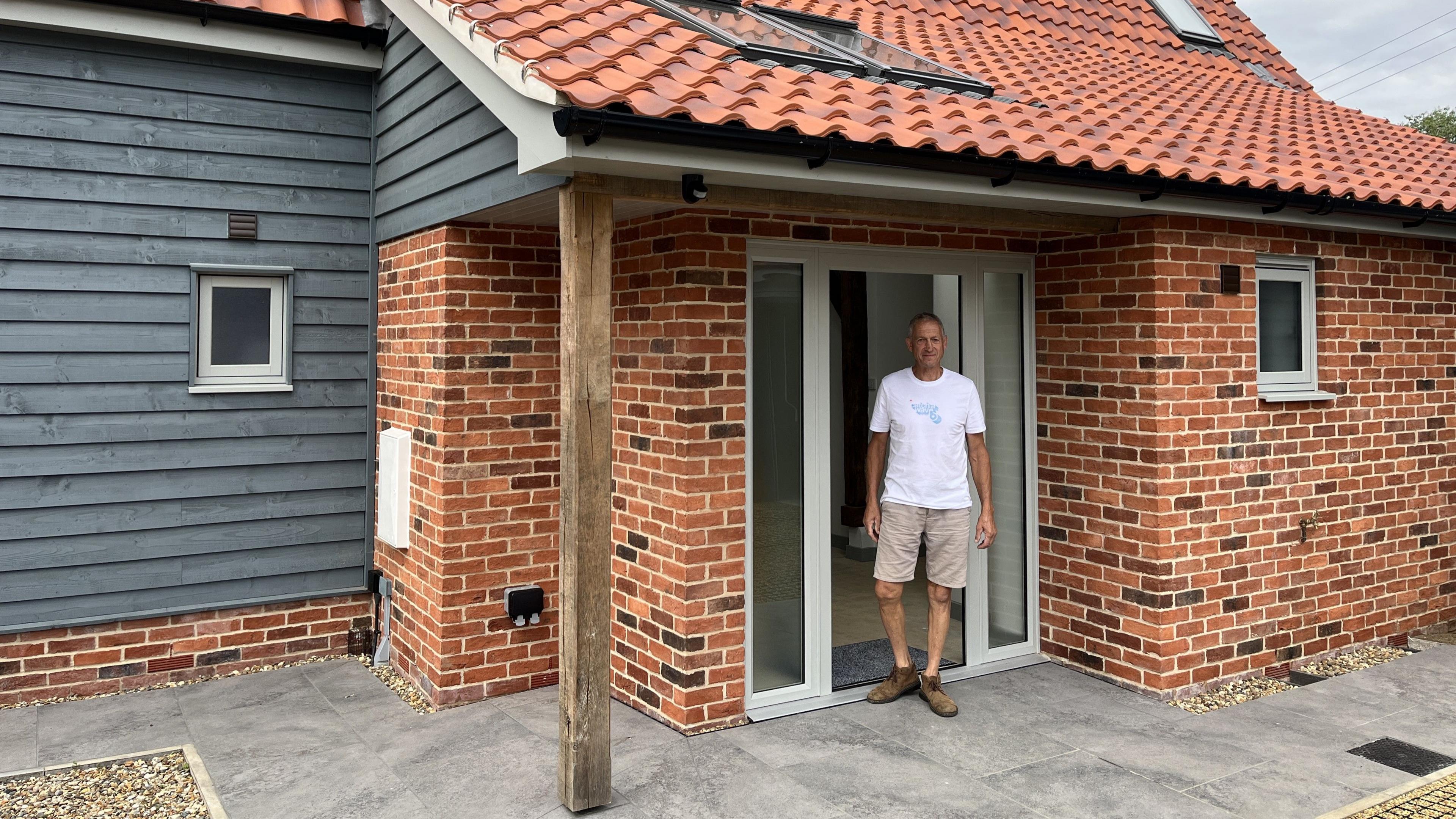 Mr Bowden smiles at the camera standing in the doorway of his home. He wears a white T-shirt with beige shorts.