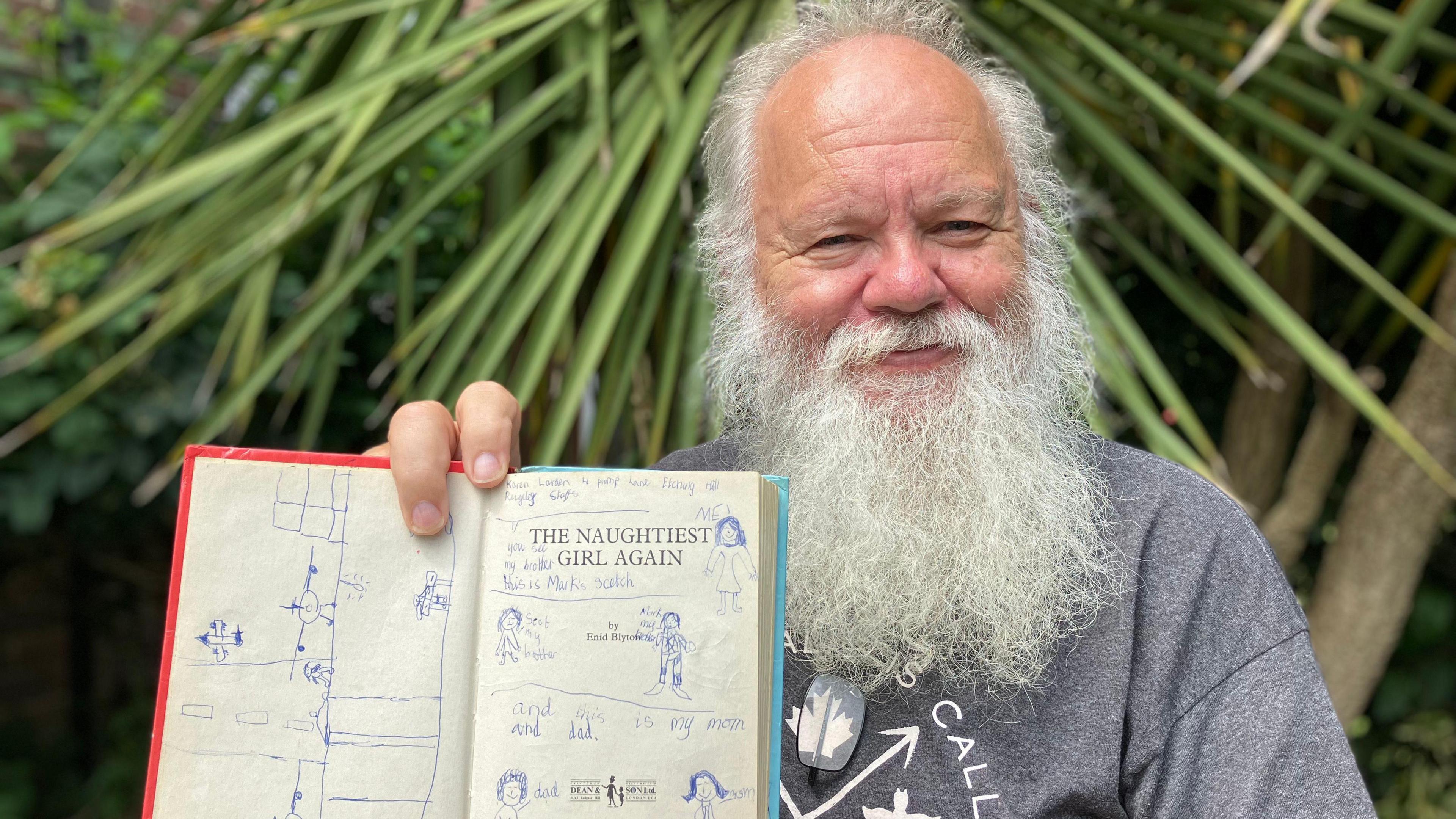 Stephen Mills has grey hair and a long beard. He is wearing a grey T-shirt and smiling while holding the open book, which has drawings of people and written notes on it. He is sitting in front of greenery.