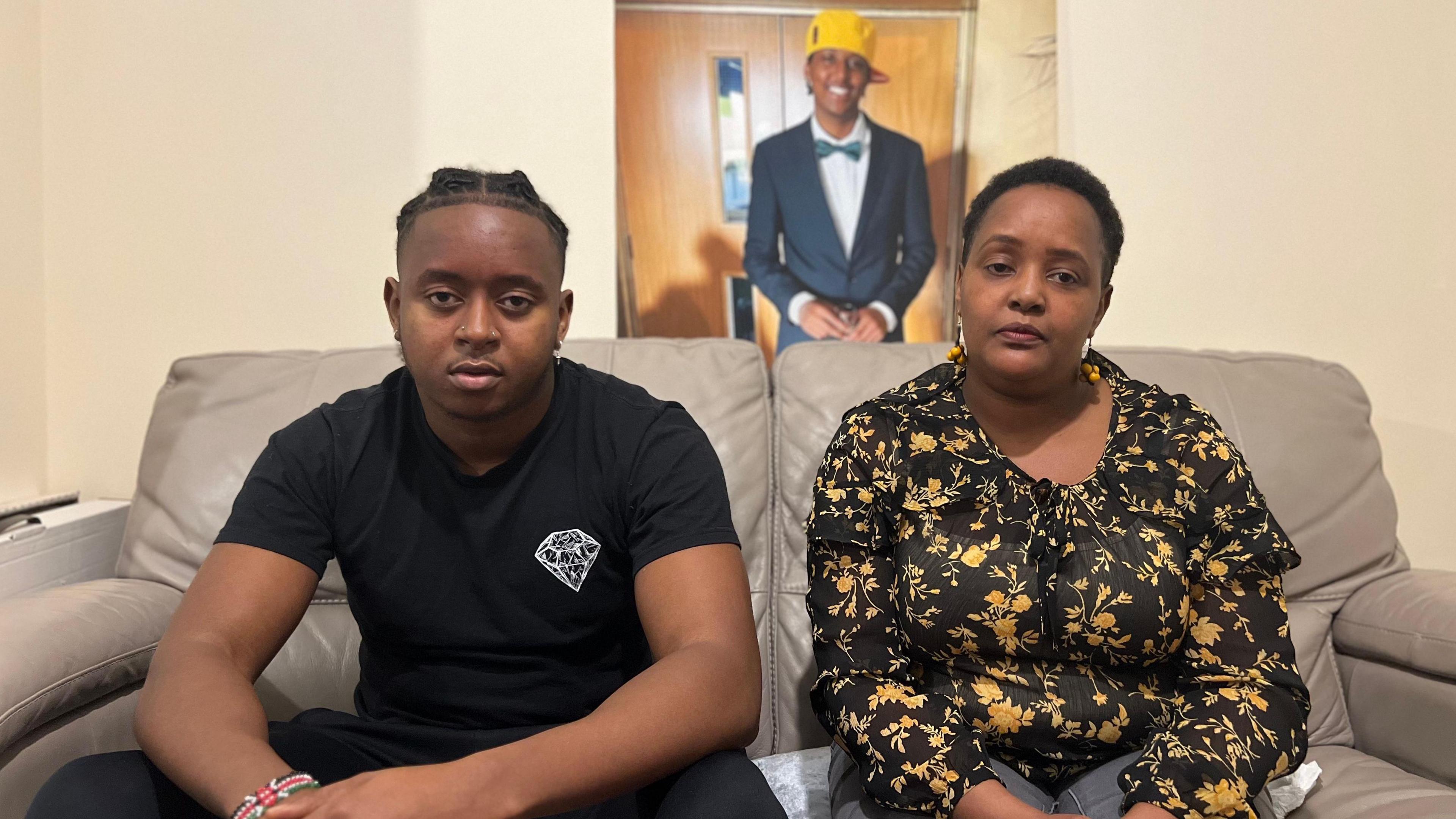 Elliot and his mum Irene are sat on a brown leather sofa. There is a photo of Eddie in the background. His is wearing a suit, a green bow tie and a yellow baseball cap 