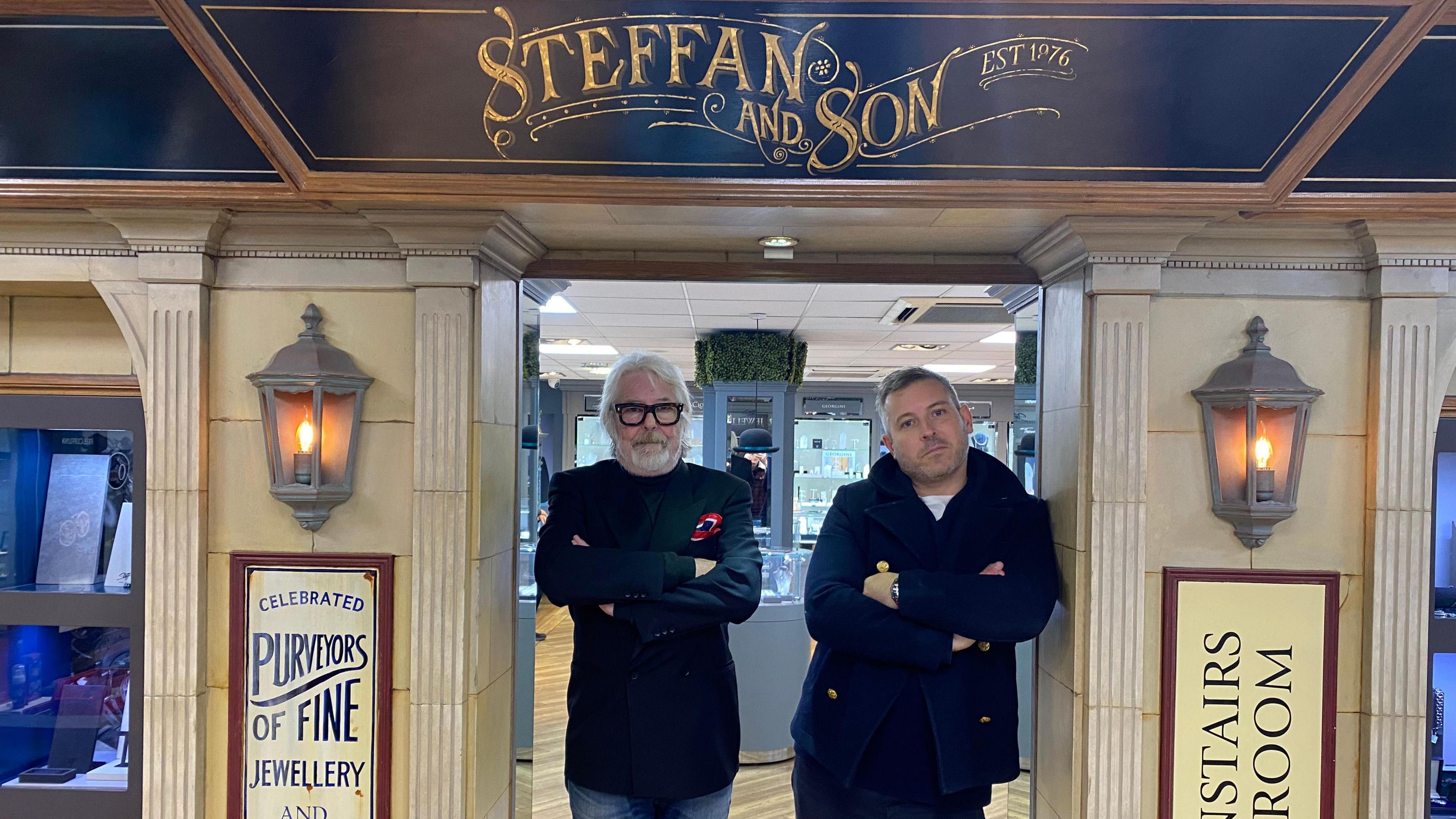 Steffan Suter stands with his arms folded in the doorway of the entrance to Steffans Jewellers, he has white hair and glasses and is wearing a navy blazer. He is standing next to his son Wes,  who has a beard and is wearing a navy coat and also has his arms folded. Neither are smiling. 