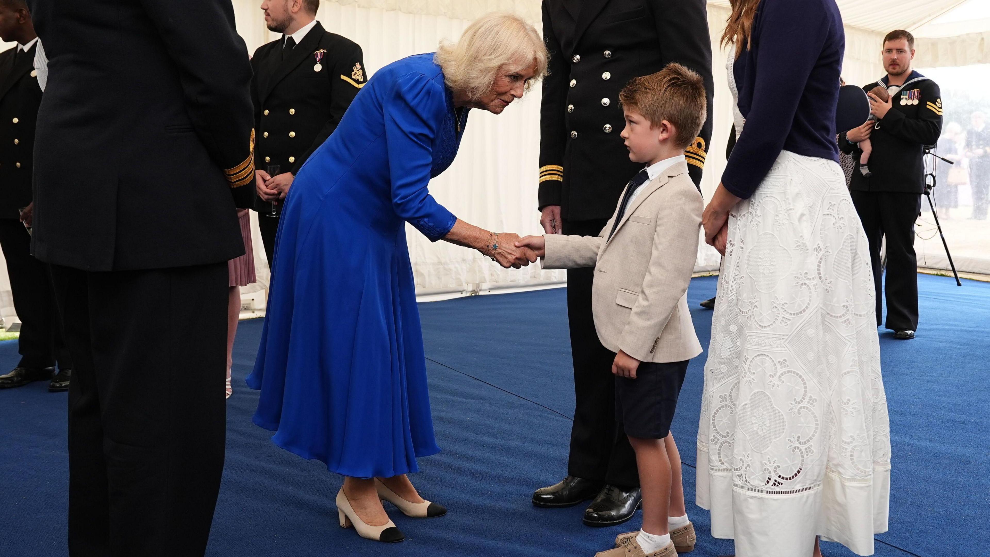 Queen Camilla, a woman with shoulder-length grey hair in a blue dress, kneels down to shake the hand of a young boy, next to his father - a middle-aged man in a navy uniform and mother - who is wearing a white dress.