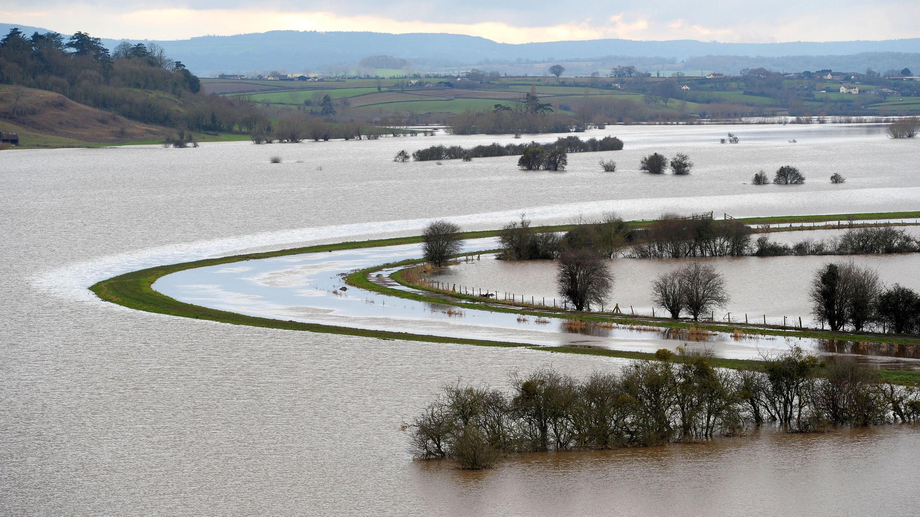 An image taken from above of extensive flooding on the Somerset Levels in 2014. A river can be seen going from left to right across the image and either side the fields are covered in water. A range of hills is visible in the distance