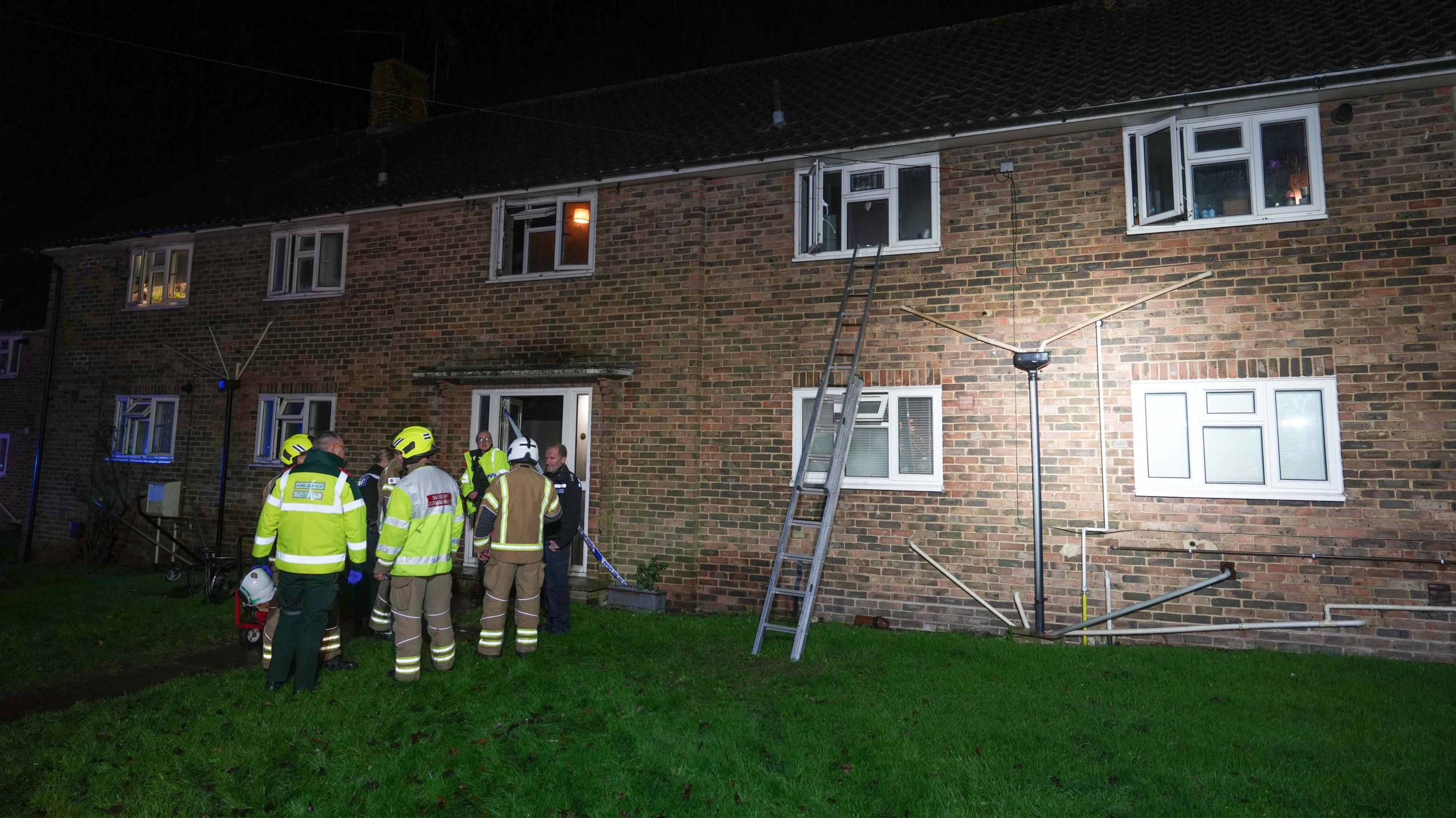 Several fire fighters in high vis stand in front of a brown brick modern building, with a ladder reaching up to a second storey window.