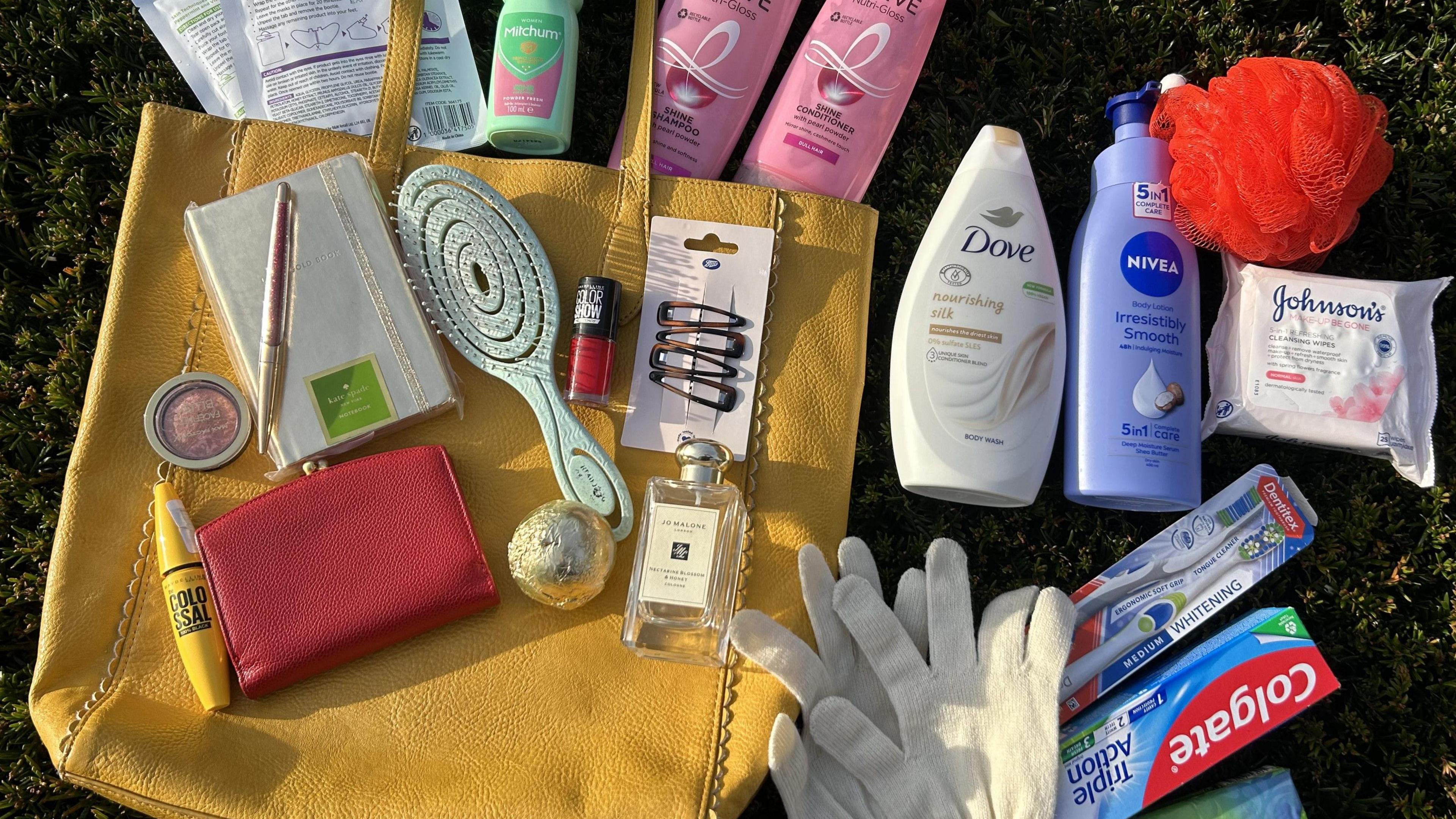 A yellow handbag laid on some grass with unopened items around it, including a toothbrush, toothpaste, gloves, mascara, perfume, purse, hairbrush, shampoo, conditioner, a notepad and pen, hair clips, make-up remover wipes and deodorant. 