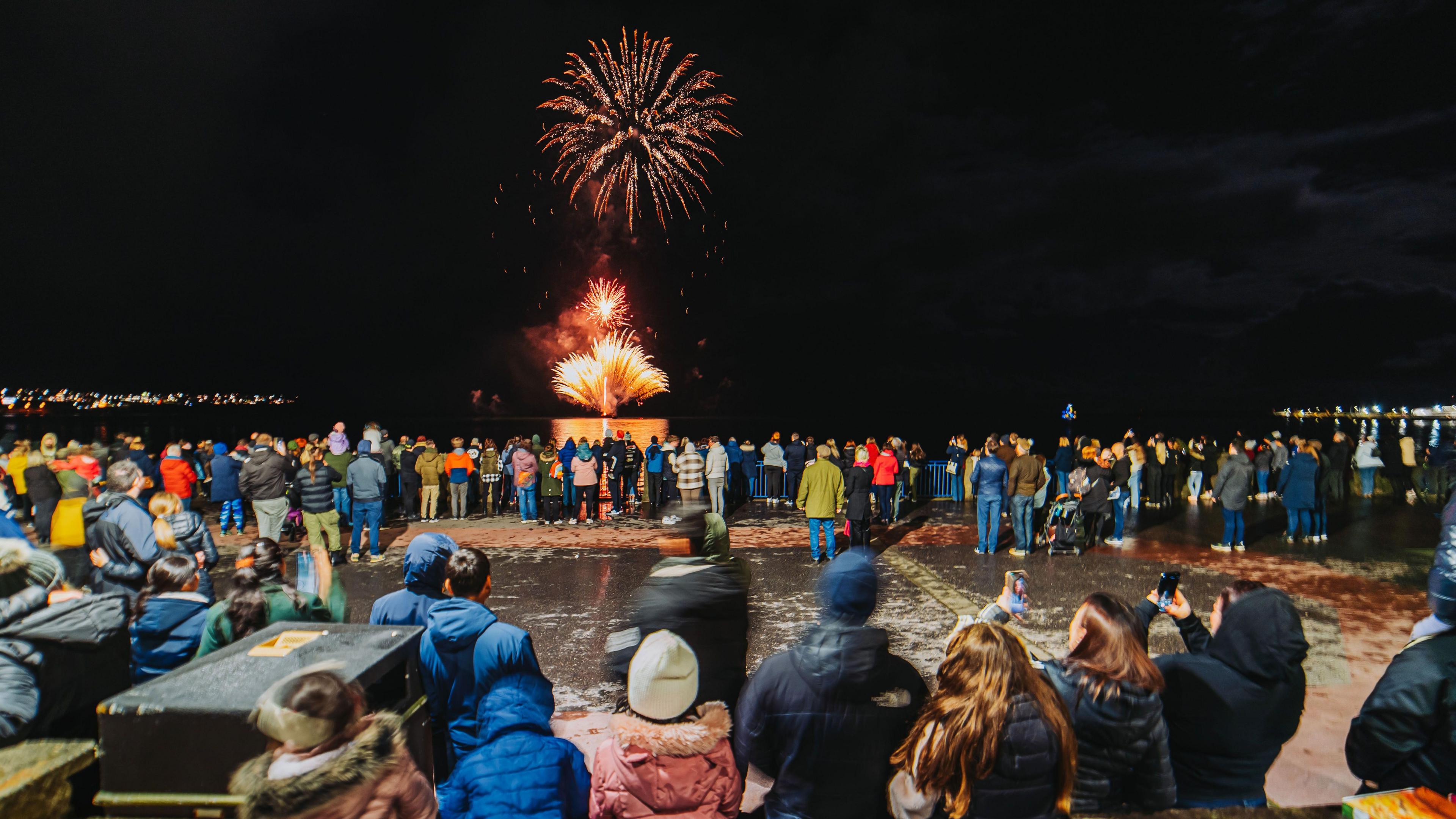 Crowds of people wearing warm winter clothes watching on as a large red firework explodes over Douglas Bay.