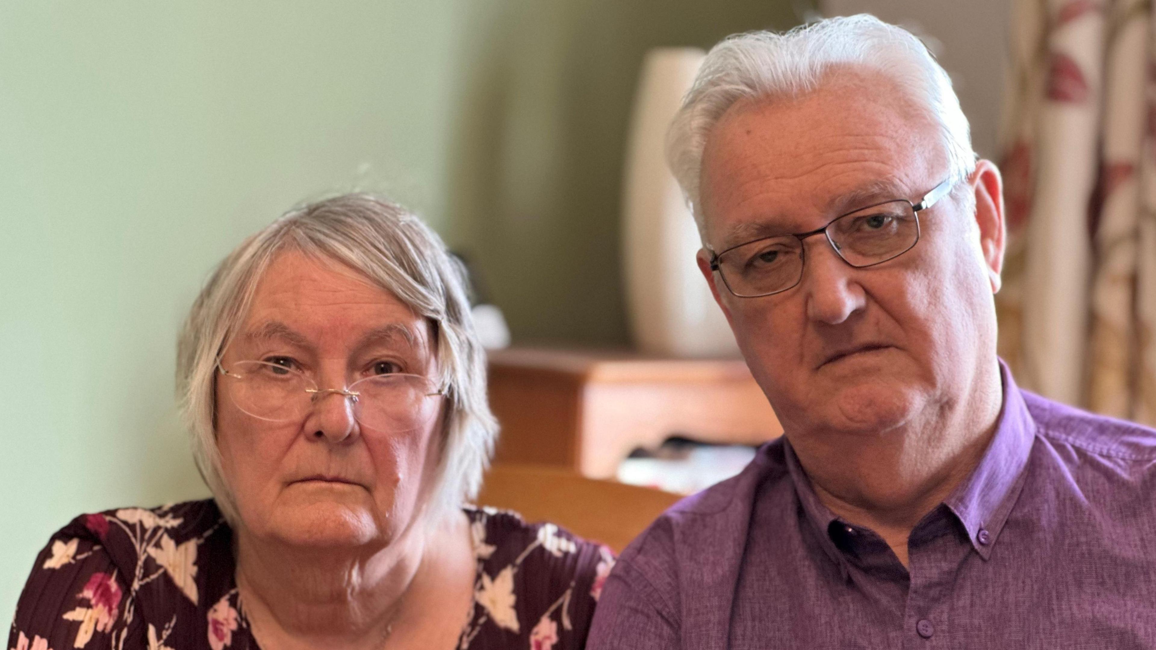 Katie and Peter McCombe are pictured looking disappointed and annoyed in the dining room of their home. They both have grey hair and are wearing glasses. The walls behind them are green and they have flowered curtains to their left. There is cabinet blurred in the background with a vase on top. Mr McCombe is wearing a mauve shirt. His wife is wearing a black and floral top.