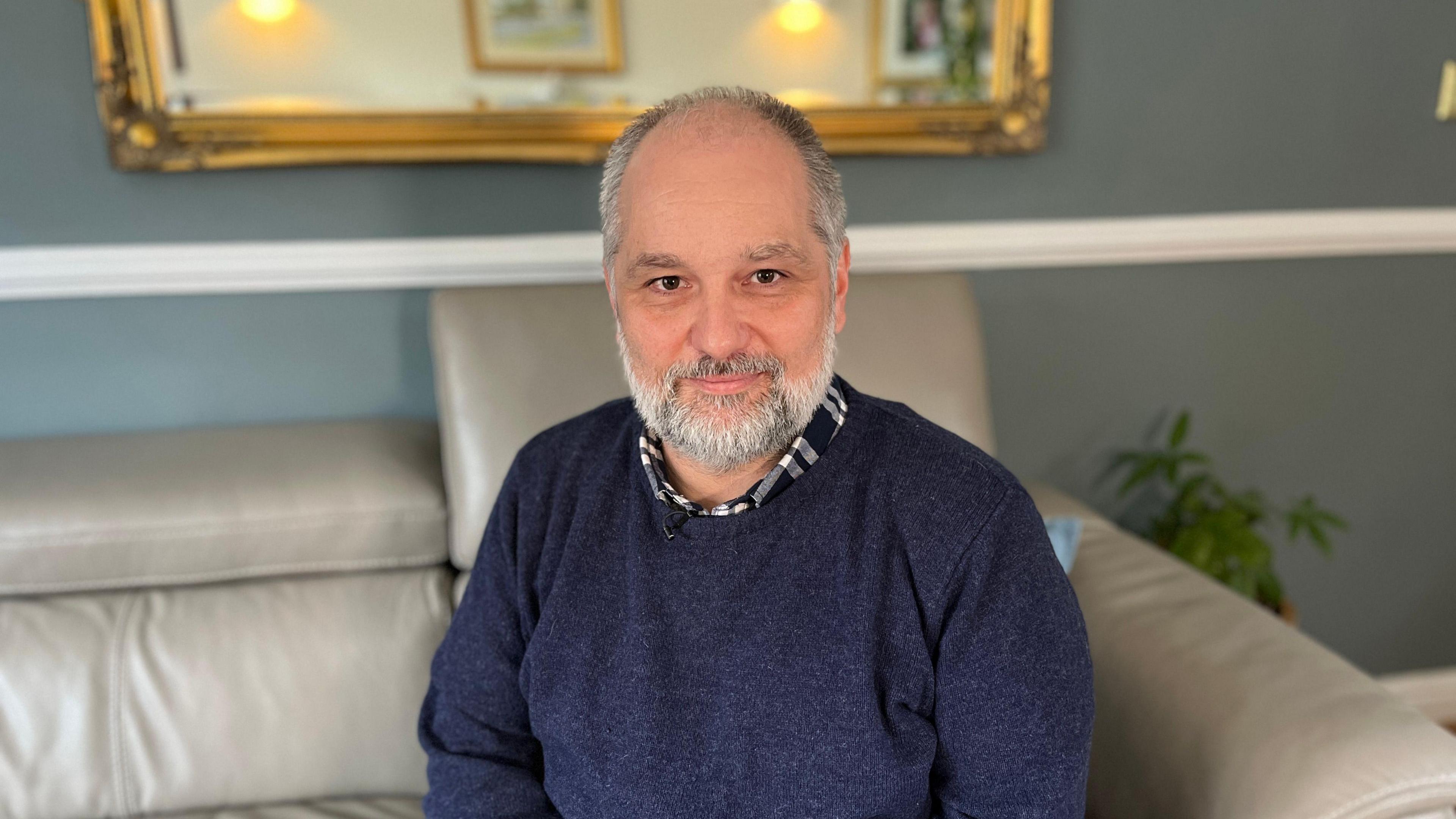 Paul White wearing a navy crew neck jumper and checked shirt, sat on a grey leather sofa