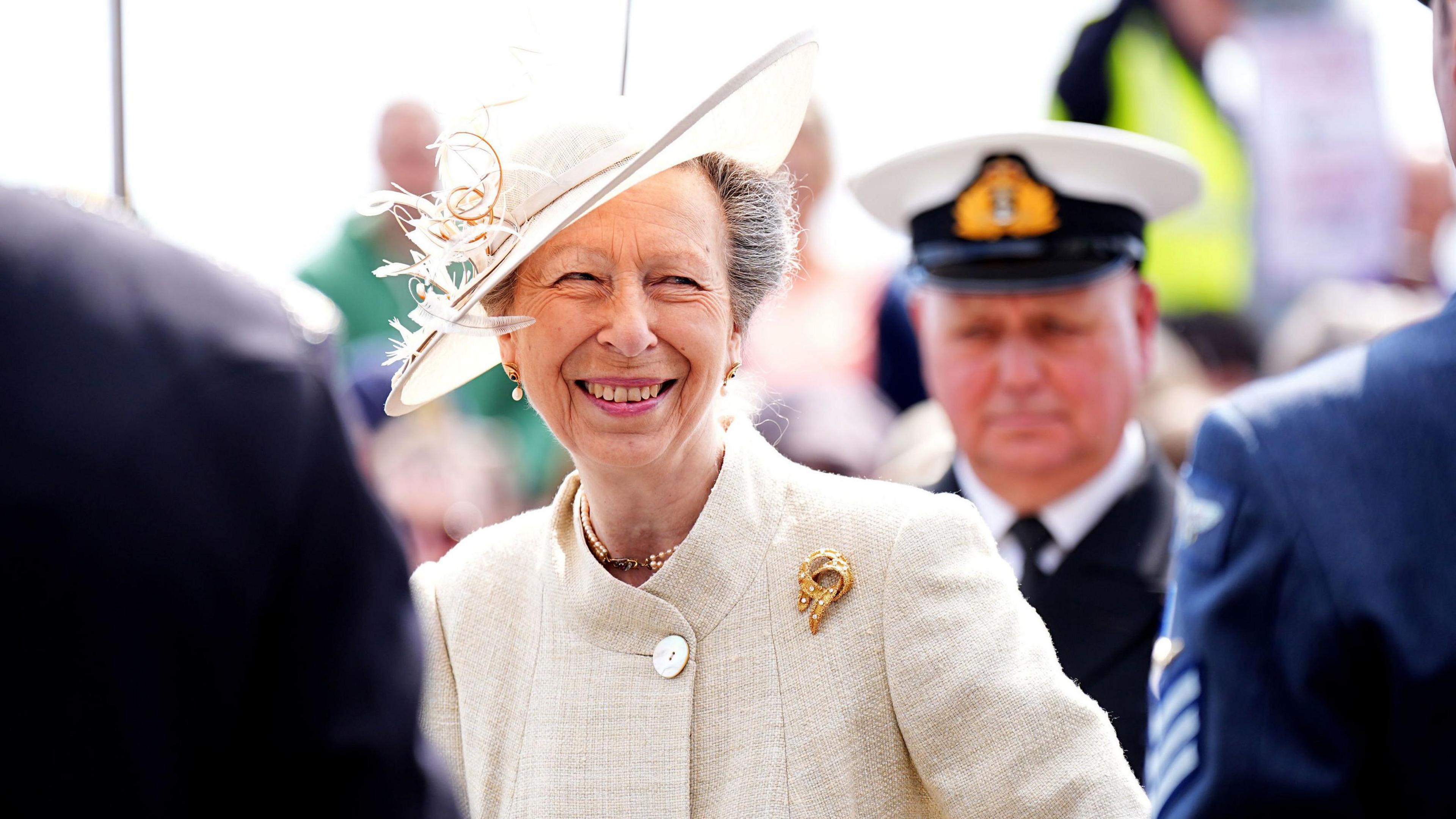 A picture of Anne wearing a white jacket and a matching hat. She has grey hair and is smiling
