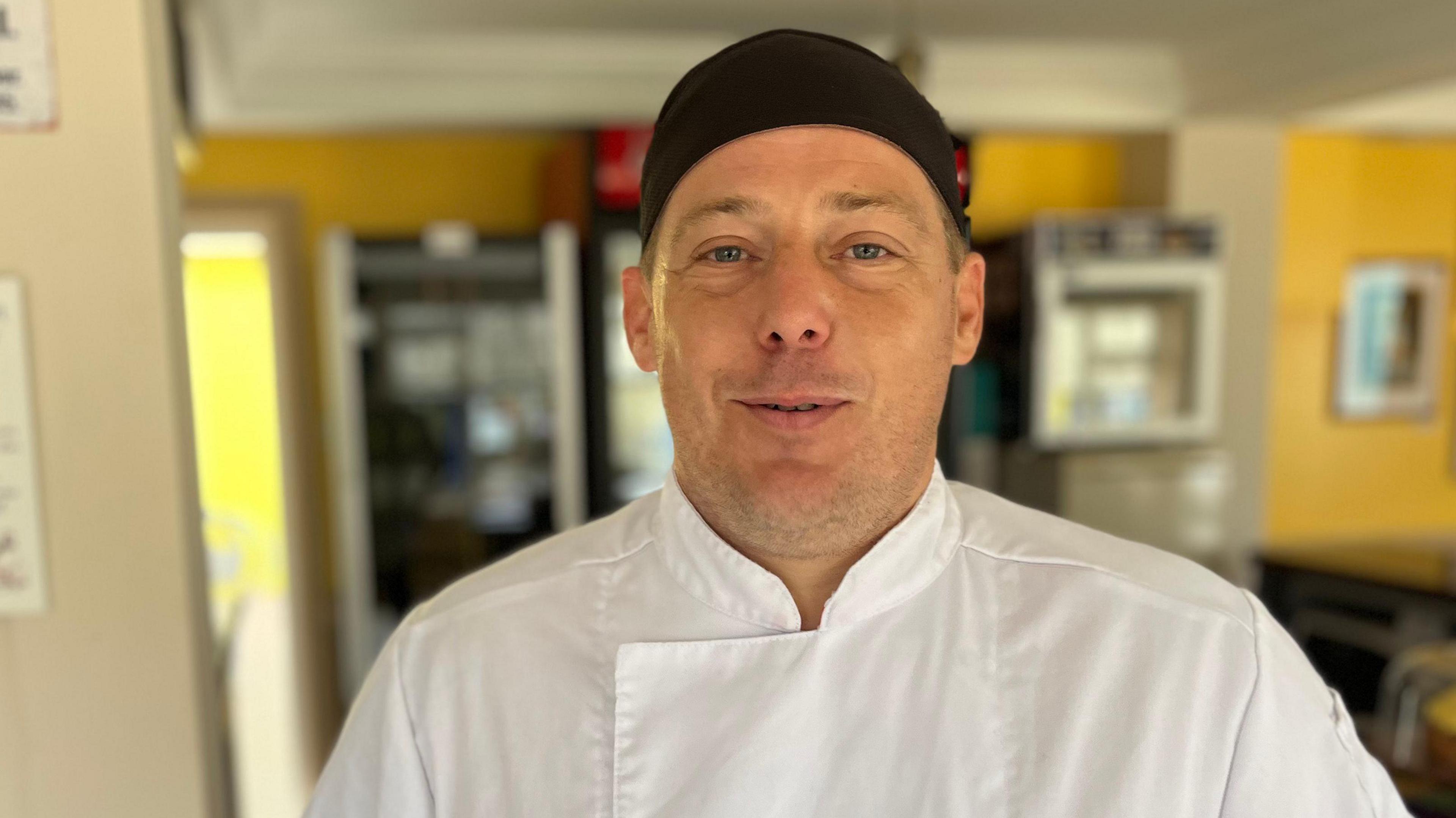 Aidan Le Poidevin, wearing chefs' whites with a black hat, looking towards camera