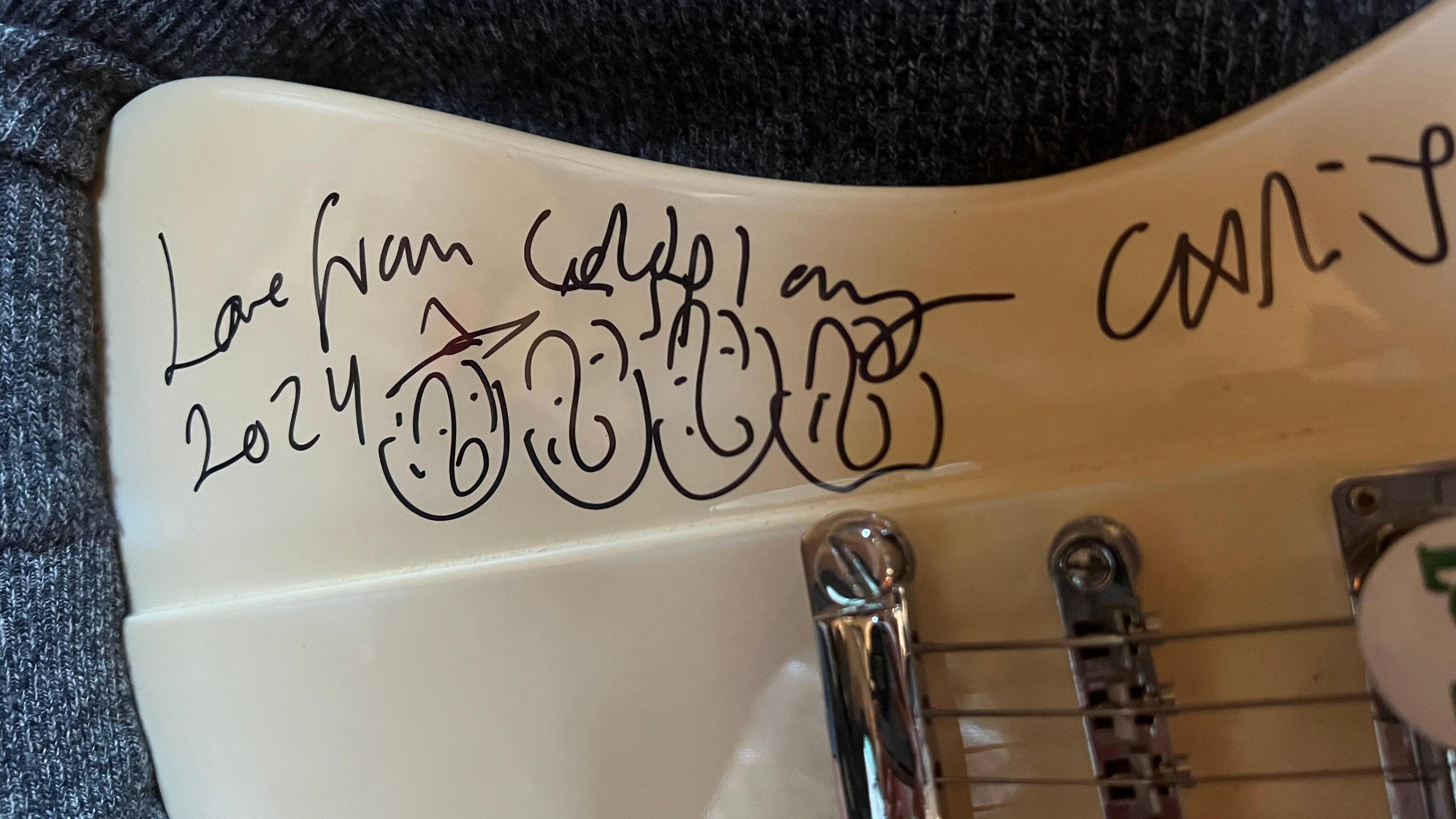 A white electric guitar is signed by Coldplay in black marker. It says Love from Coldplay 2024. There is a drawing of four faces next to it.