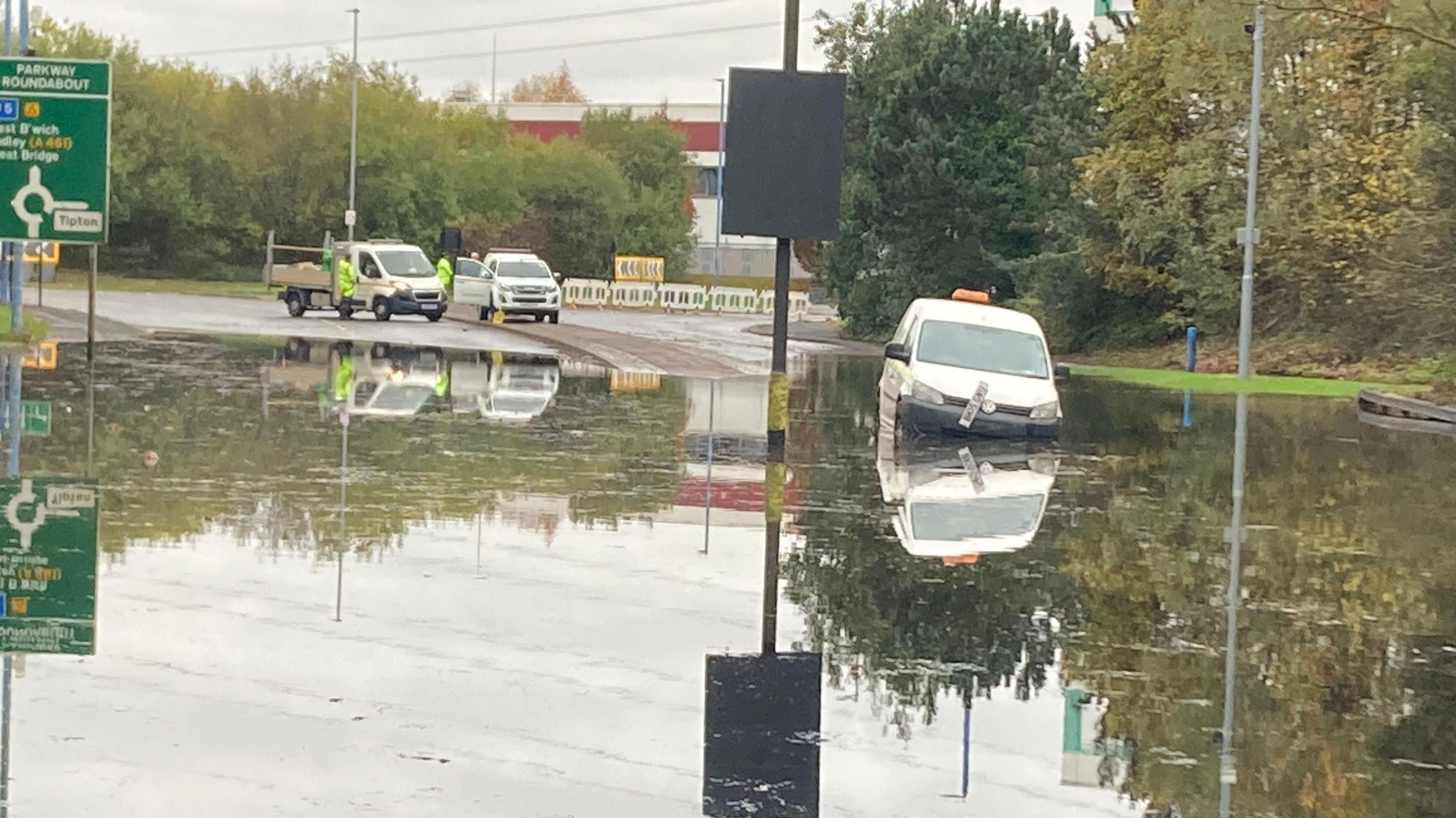 A view of a flooded road with a white van parked with water up to its bumper and a broken number plate 