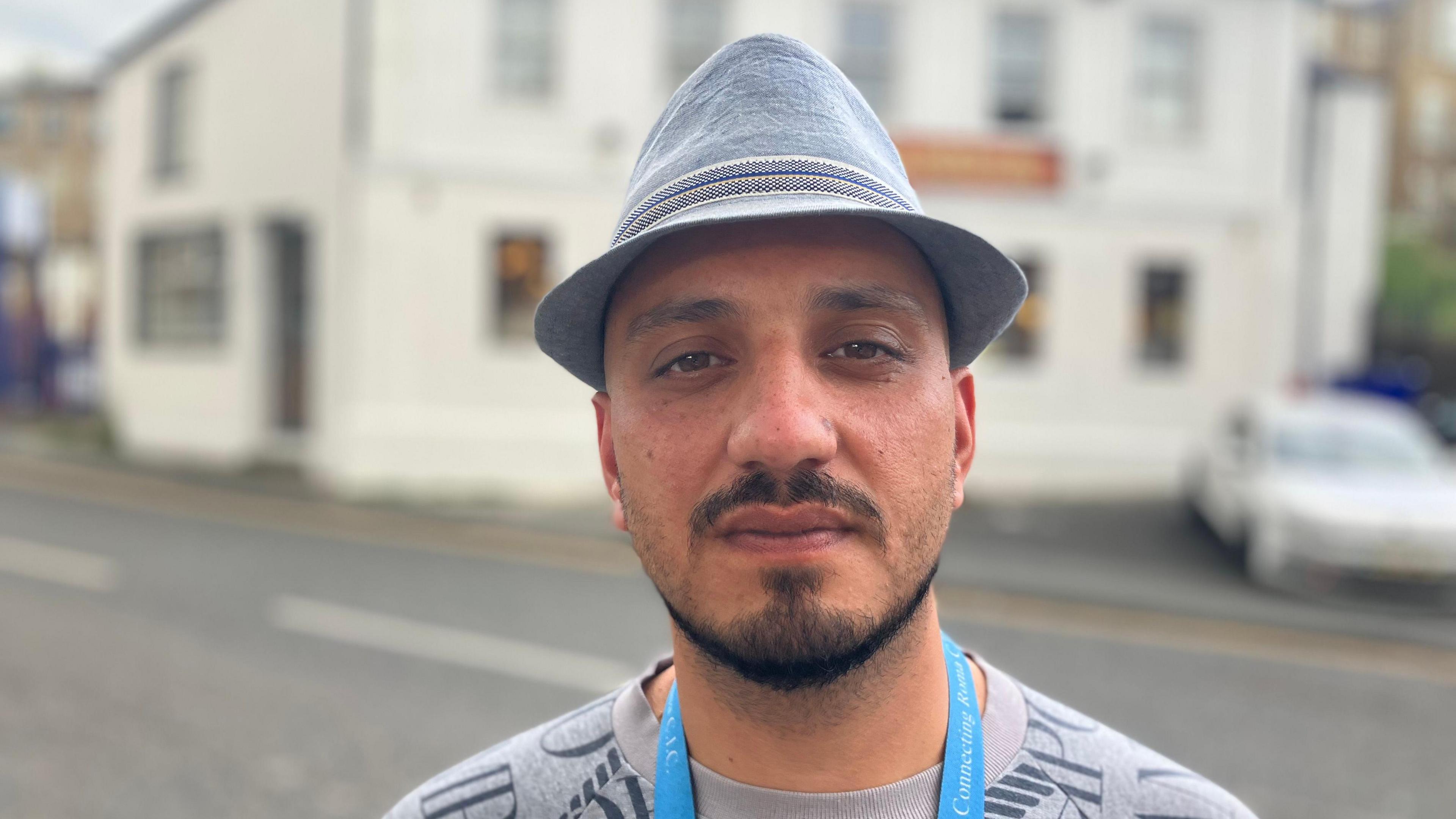 A man with a black beard looks at the camera. He wears a grey trilby and a grey top. A blue lanyard hangs around his neck.