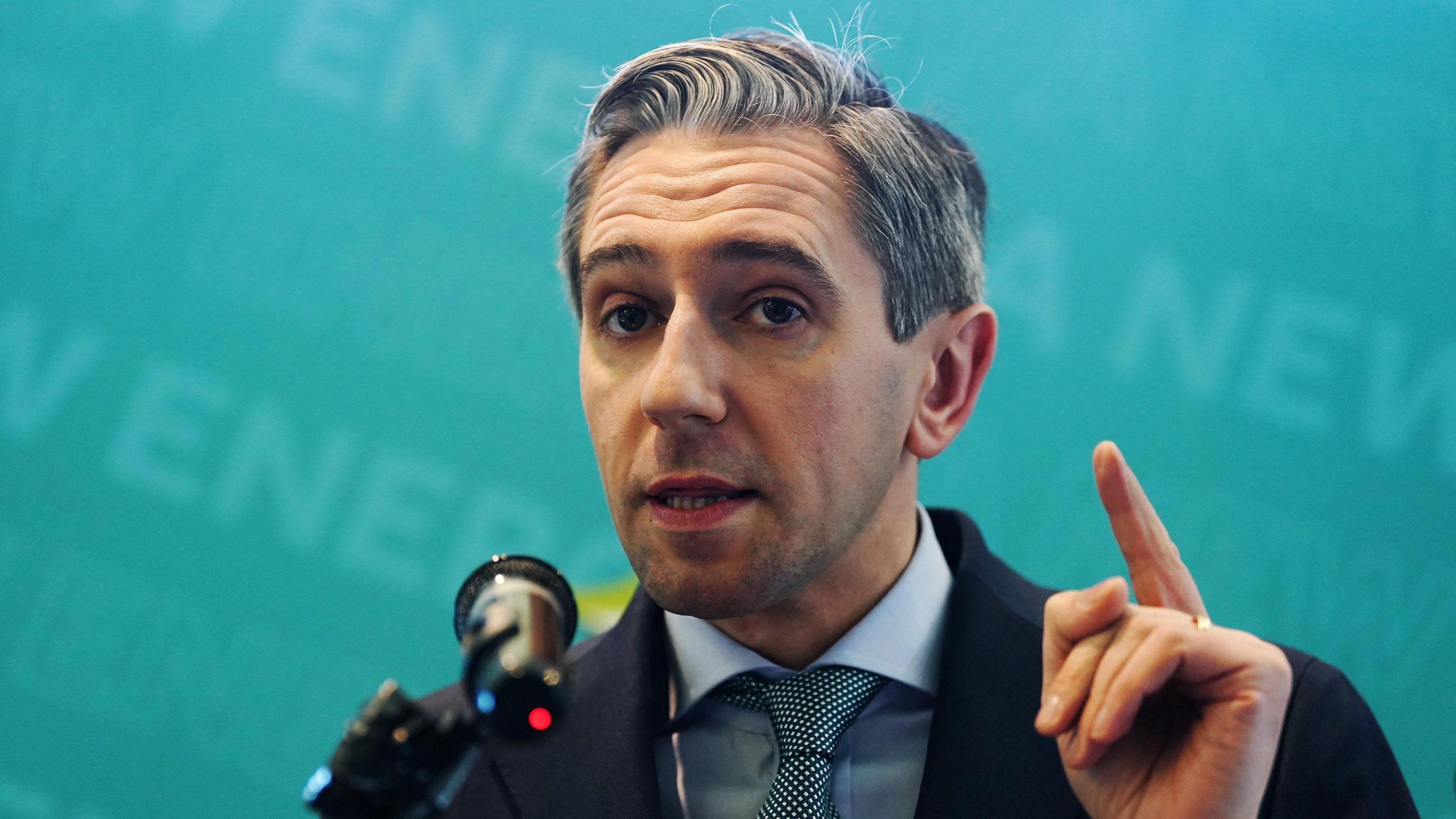 The Taoiseach Simon Harris delivering a speech. He is wearing a navy and blue suit. He has a finger in the air. 