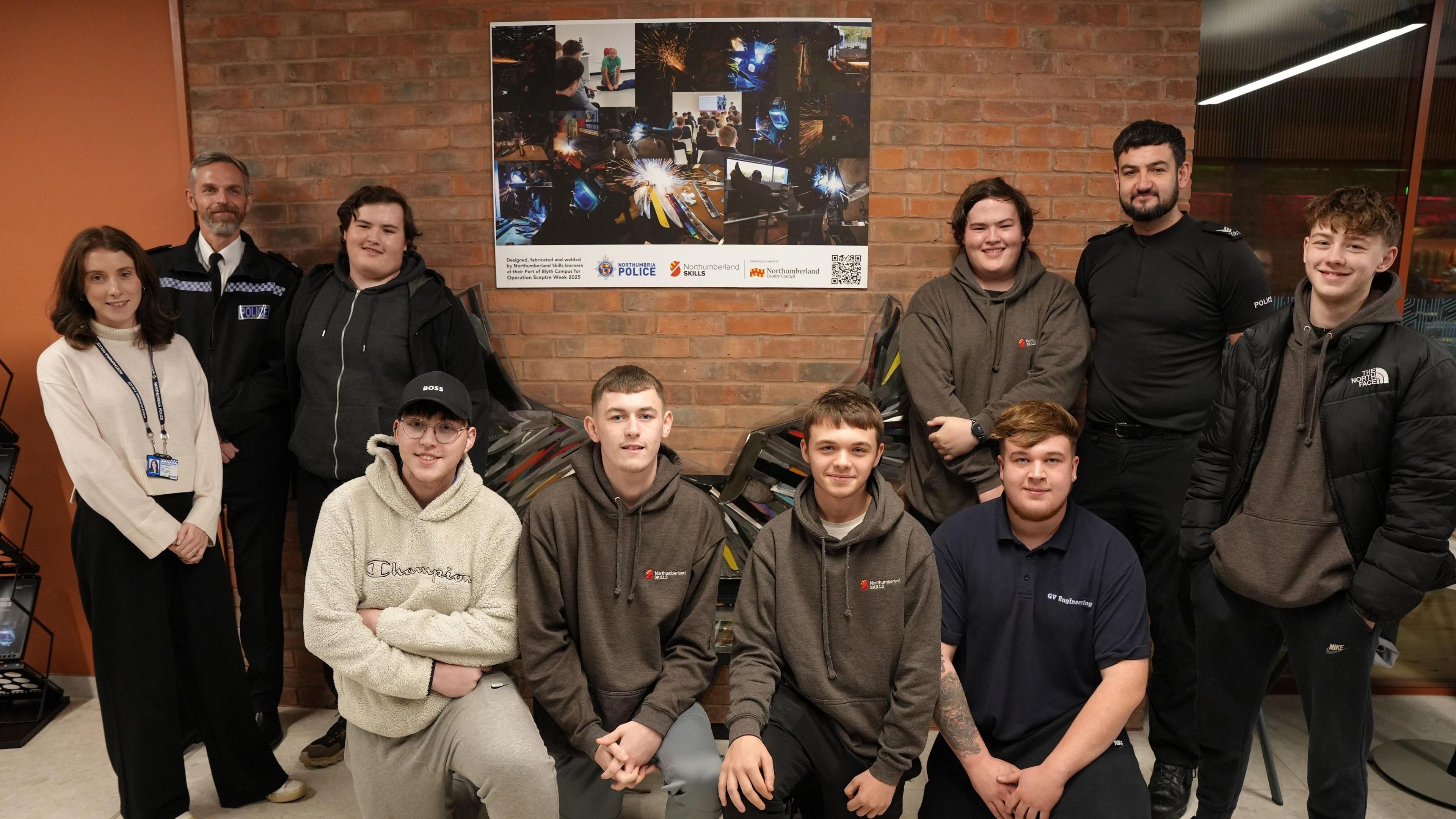 A group of students are gathered around the bench, with four young men kneeling in front of it, and three students to the right of it. To the left of the bench is one other student as well as Superintendent Scott Cowie and a female member of staff.