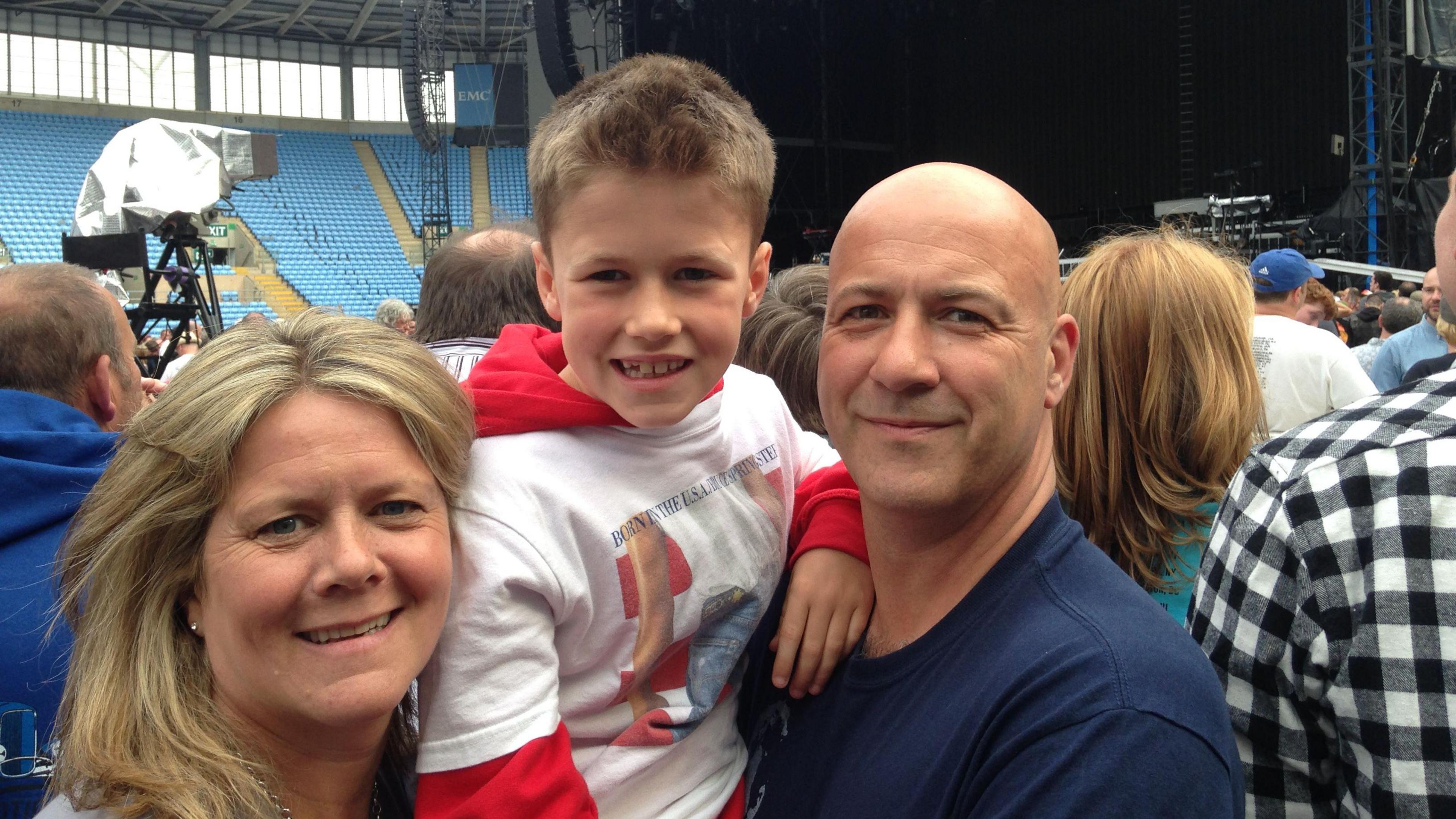 A young boy smiles at the camera. He has a red hoody on and a white t-shirt over the top. He is being held up by a man who has a blue t-shirt and a bald head who is also looking at the camera. On the other side of him is a woman with blonde hair and a grey t-shirt. They are all cuddled up posing for a photo, behind them are fans in a stadium all facing towards the stage, which can be seen at the back of the photo.