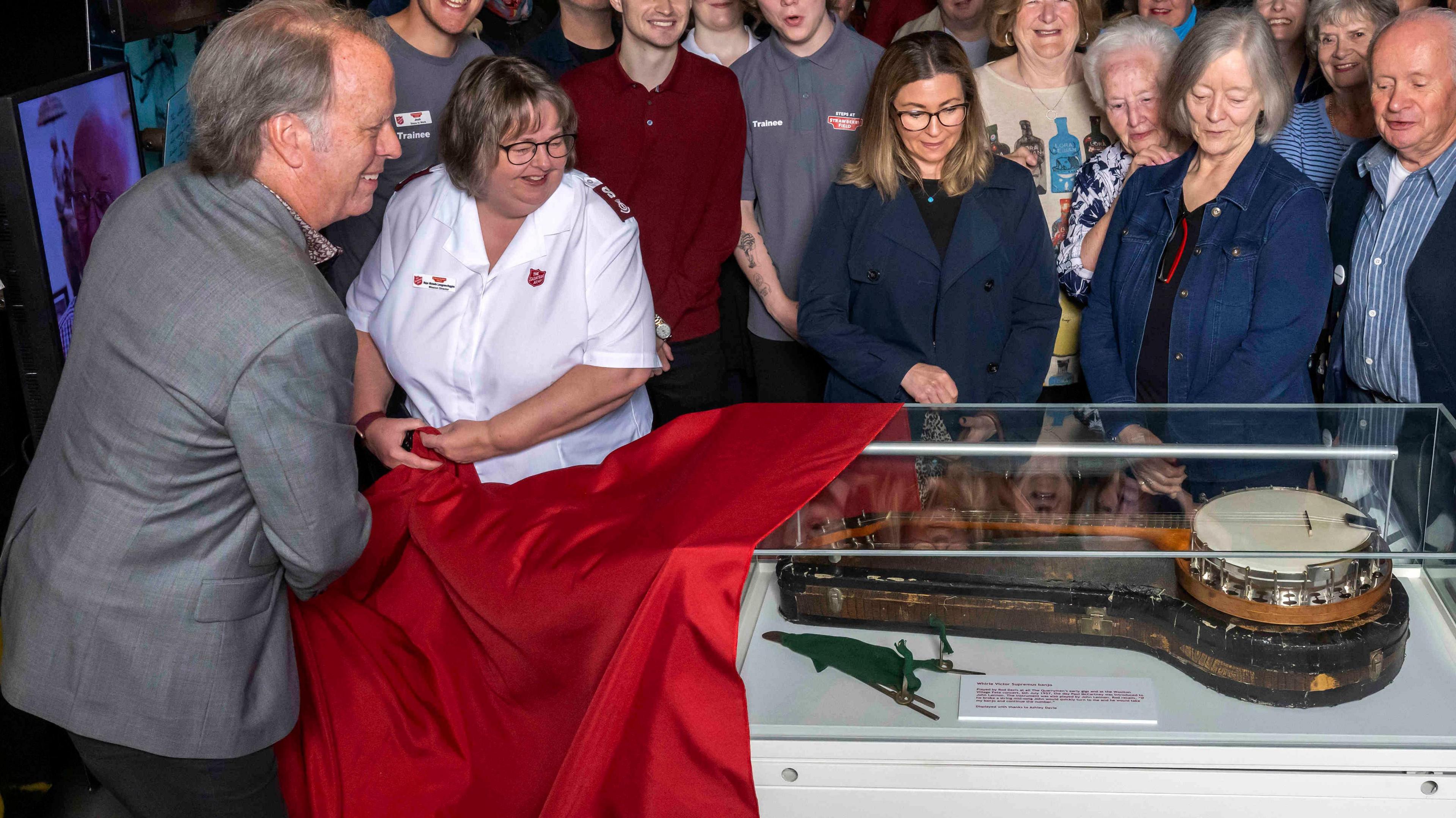 A man in a grey jacket and a woman in a white uniform pull a red cloth off an exhibition case, which holds a white and black banjo once played by John Lennon