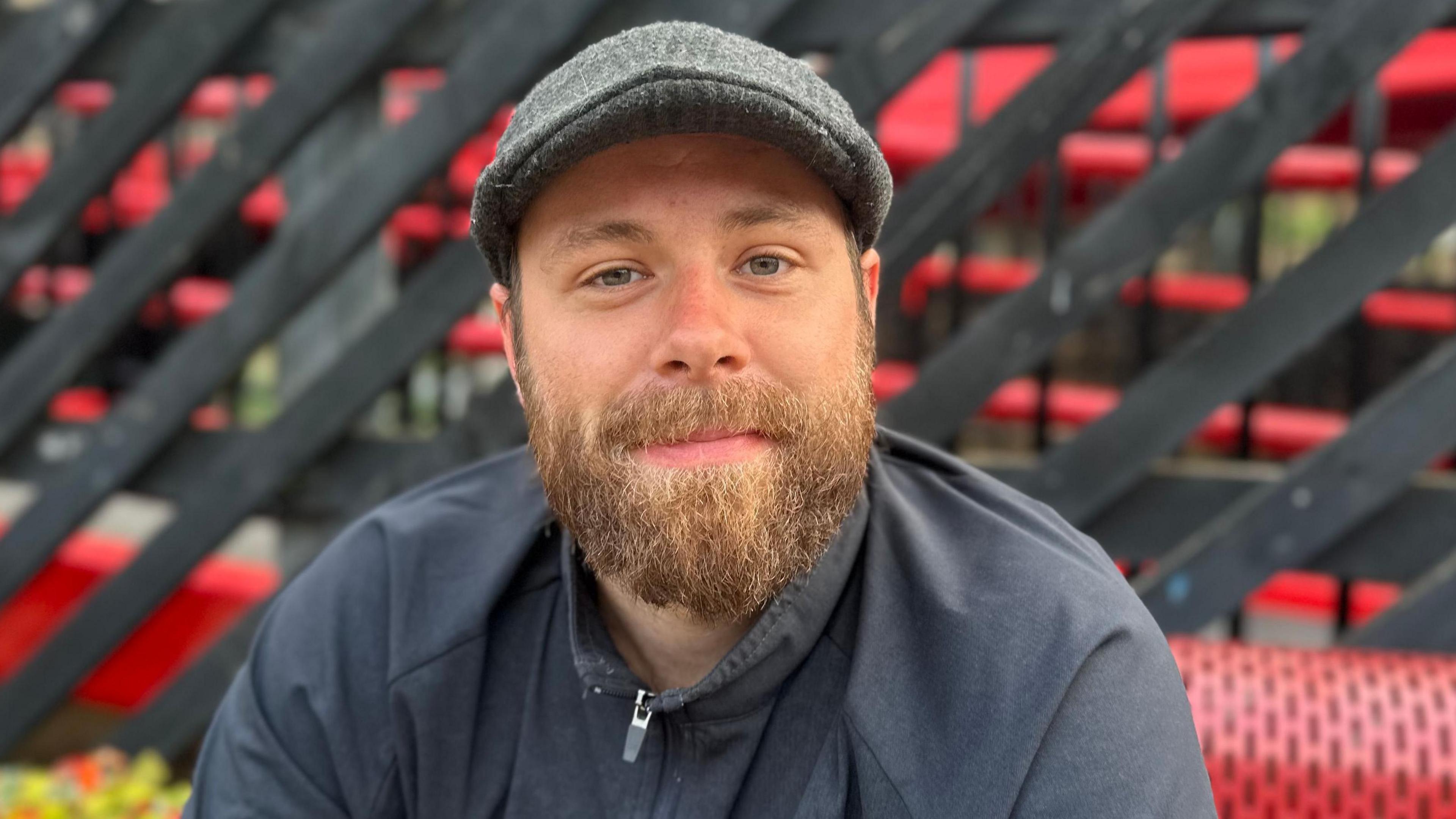 Conn-ah Norman, who is wearing a grey flat cap. He has a brown beard and is staring directly at the camera.