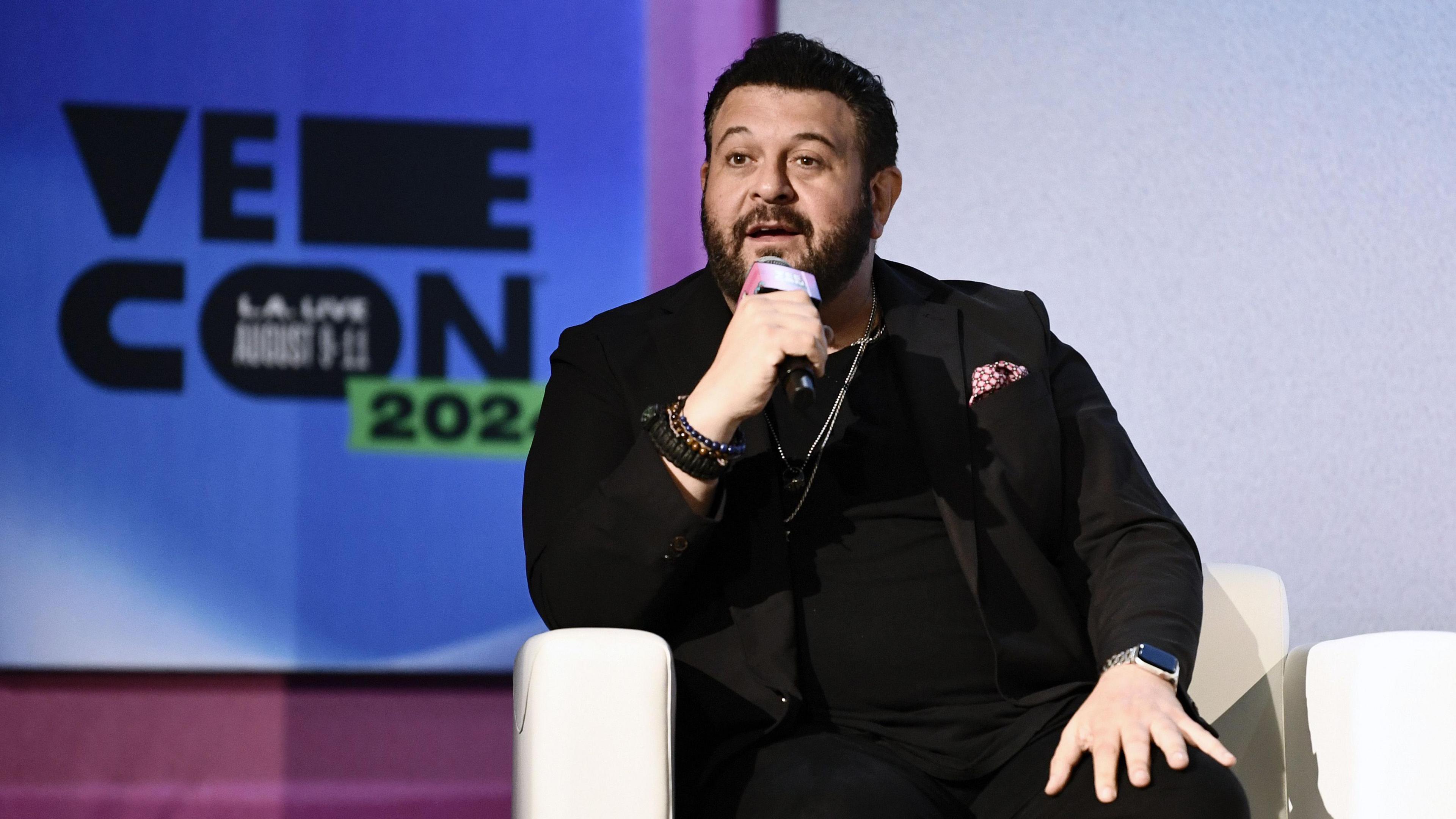 Mr Richman is sat on a white arm chair speaking through a microphone during expo VeeCon 2024, with its signage in the background. He is  wearing all black with a pink dotty pocket square and is wearing an Apple Watch. 