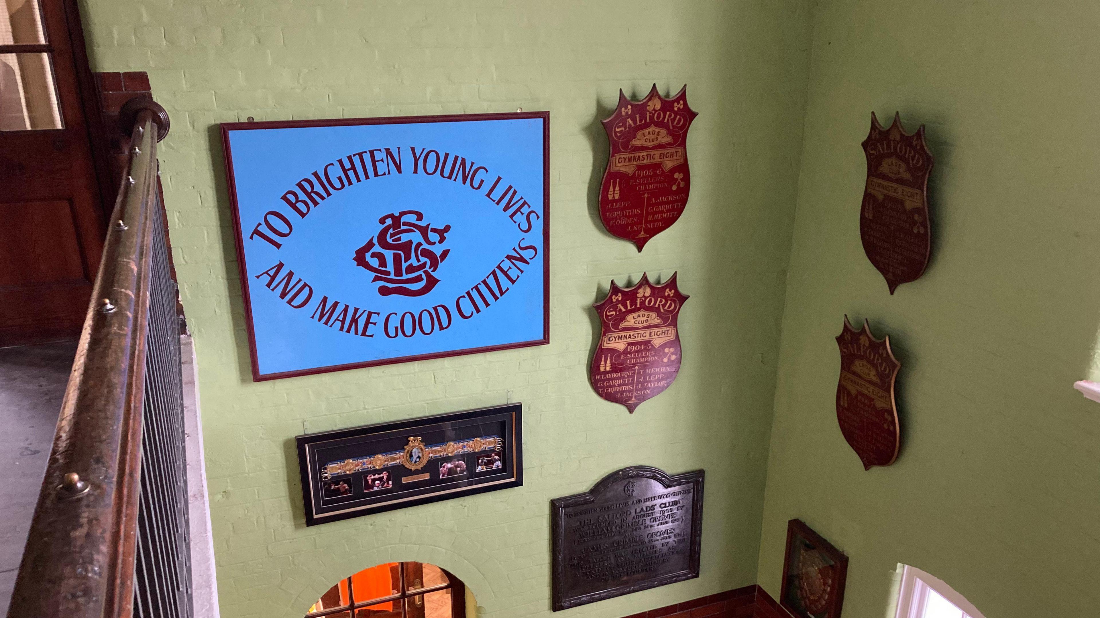 A collection of plaques and signs hung against a light-green painted wall within Salford Lad's Club. One reads 'To brighten young lives and make good citizens', which is the club's motto. 