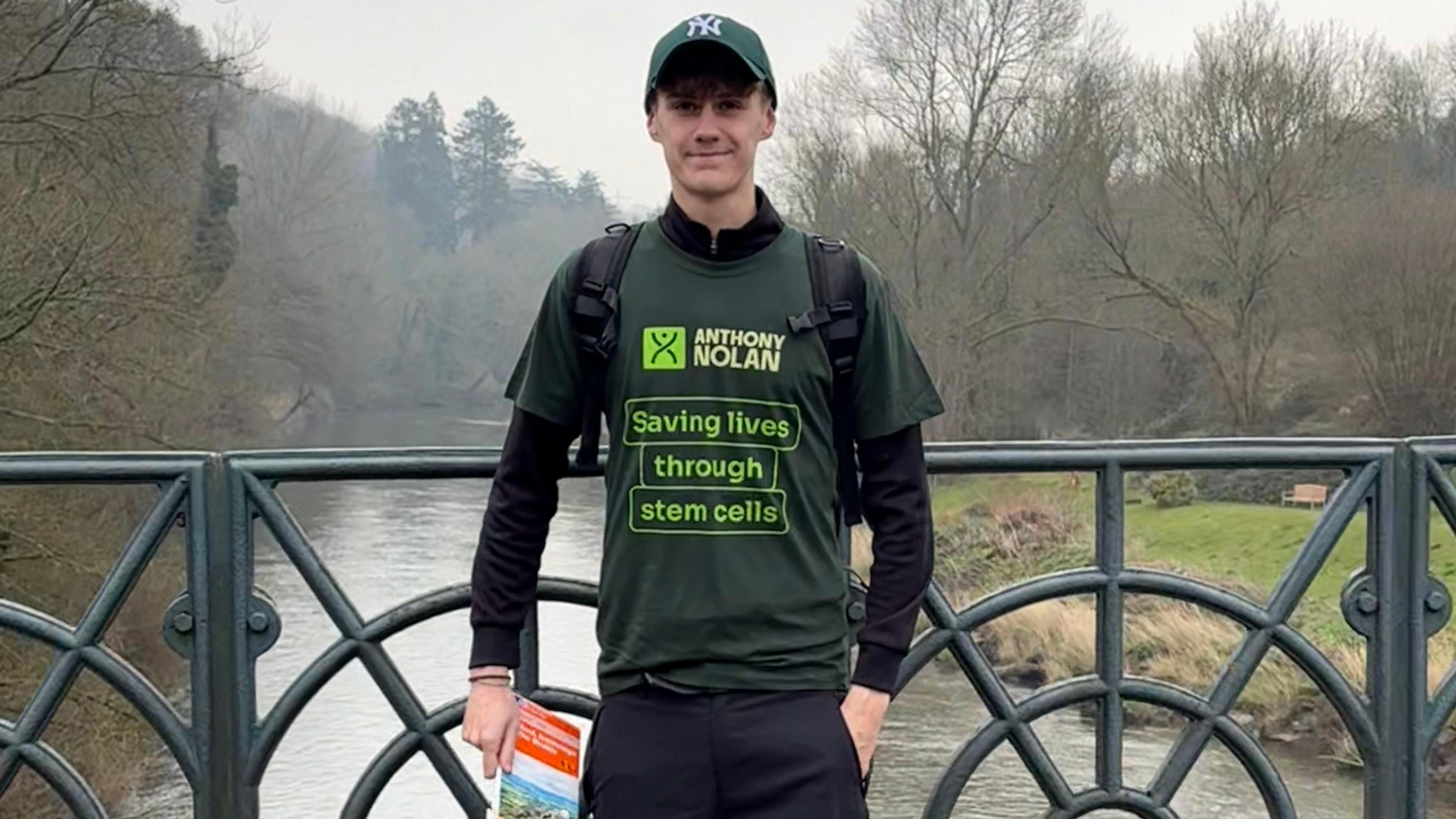A young man standing on a bridge over a river. He is wearing a green New York Yankees baseball cap and a green T-shirt which reads: "ANTHONY NOLAN - Saving lives through stem cells". He has a rucksack on his back and is holding what looks like a map.