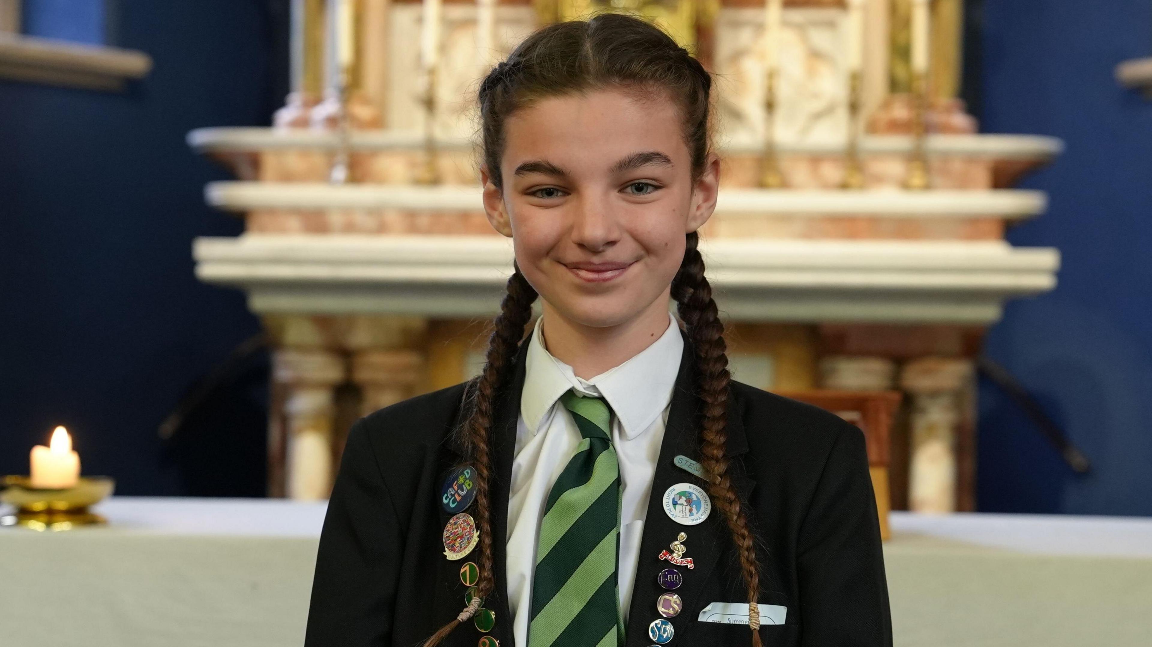 Persy wears a school blazer with multiple badges on. She has braided hair and is smiling. She wears a white shirt and a green-striped tie.
