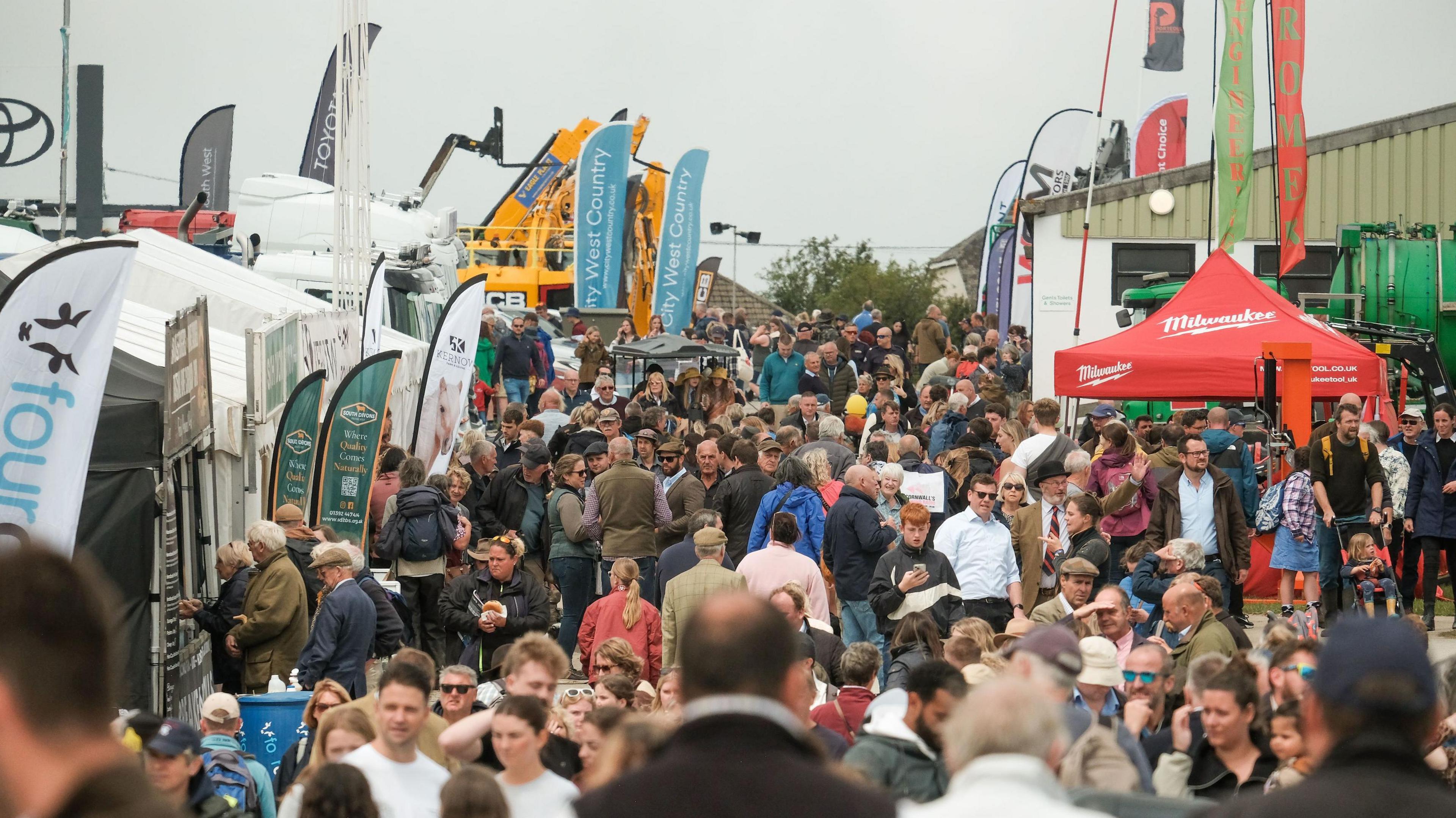 Hundreds of people at the Royal Cornwall Show which is held in Wadebridge. Stalls with large flags can been seen to either side and large yellow farm machinery in the background. 
