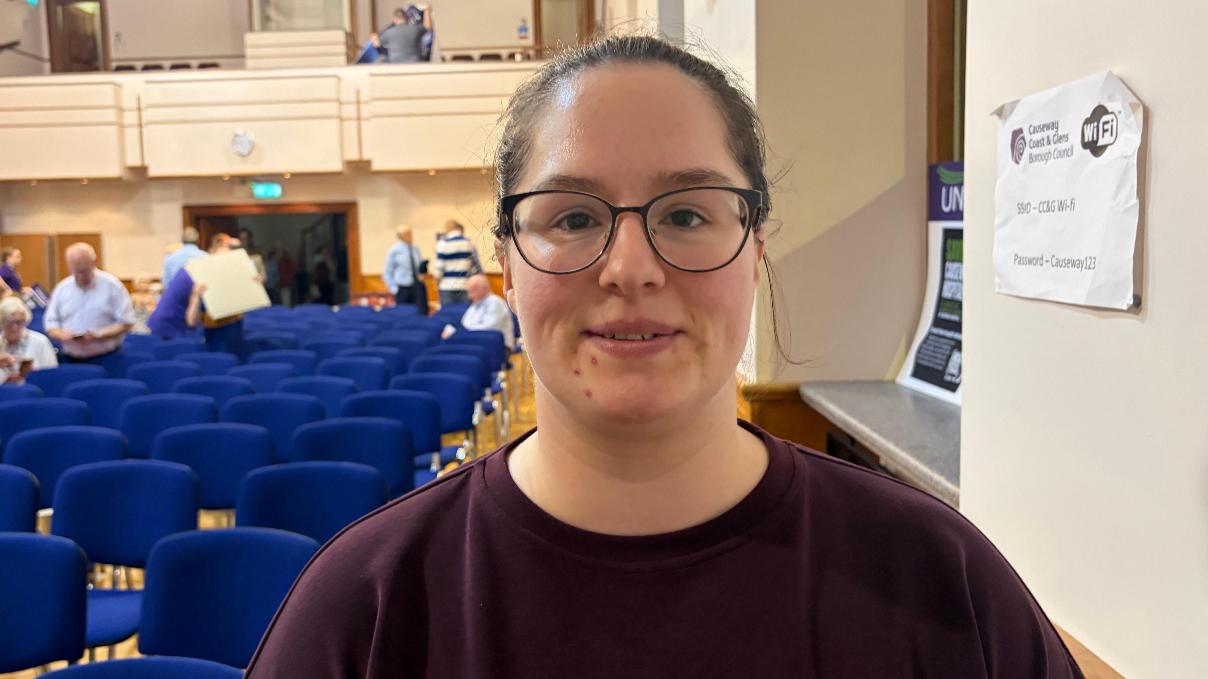 Rebecca Gill stands in Coleraine Town Hall, blue chairs can be seen behind her. She has black glasses and her brown hair is pulled back behind her head. She is wearing a maroon sweater.