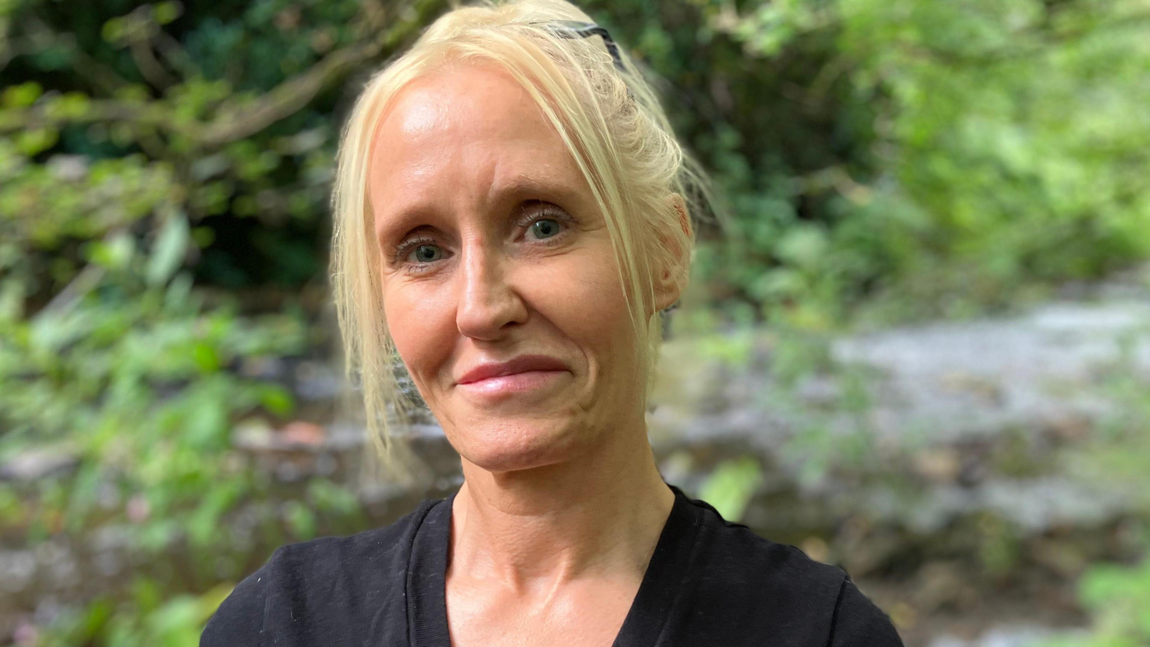 Andrea Marland is standing alongside the River Tonge in Bolton. It is a bright dry day and she is wearing a black top. Green foliage and the water can be seen in the background.