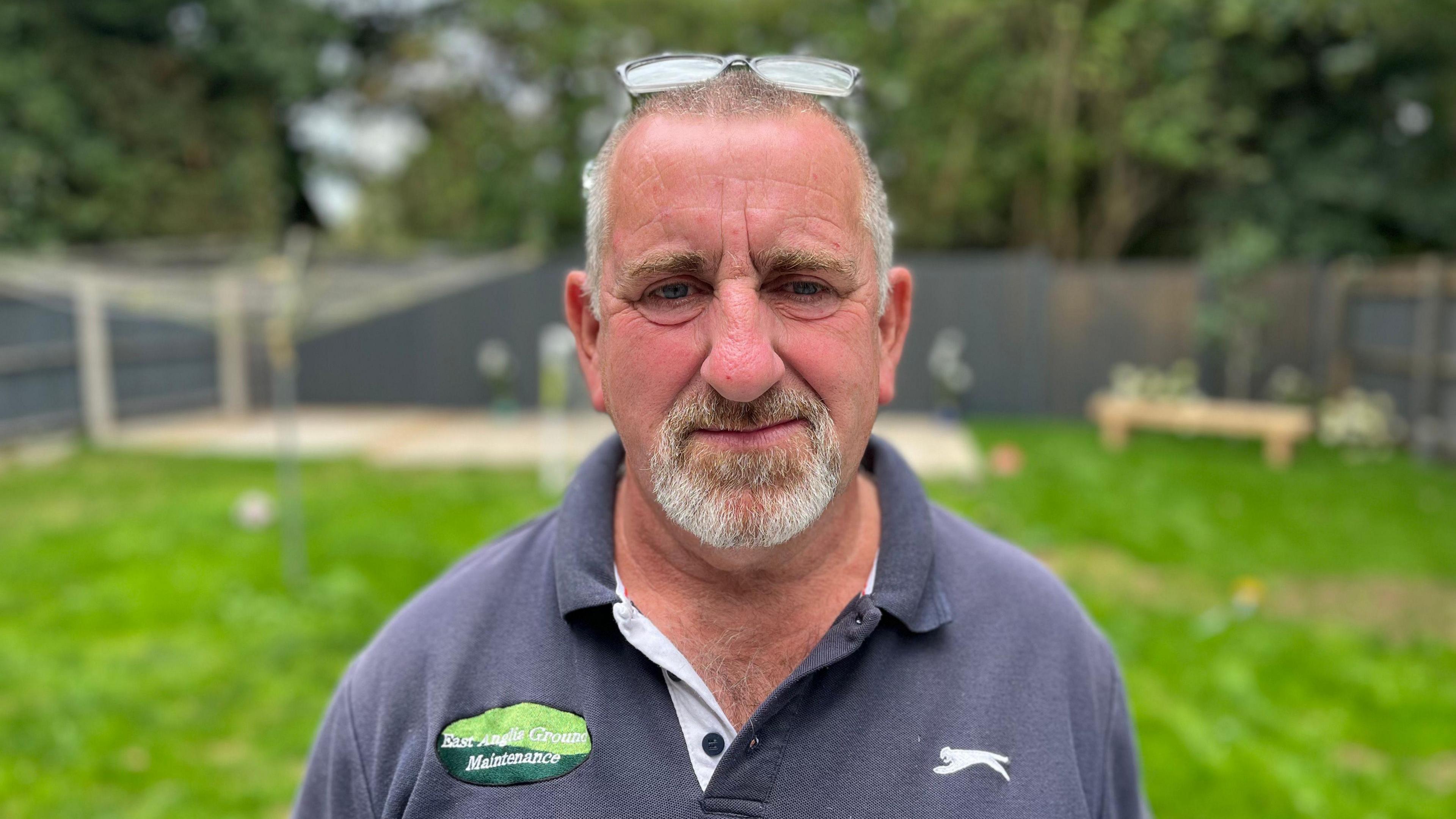 A man with a goatee beard and glasses on top of his head wearing a grey polo short faces the camera with a garden in the background.