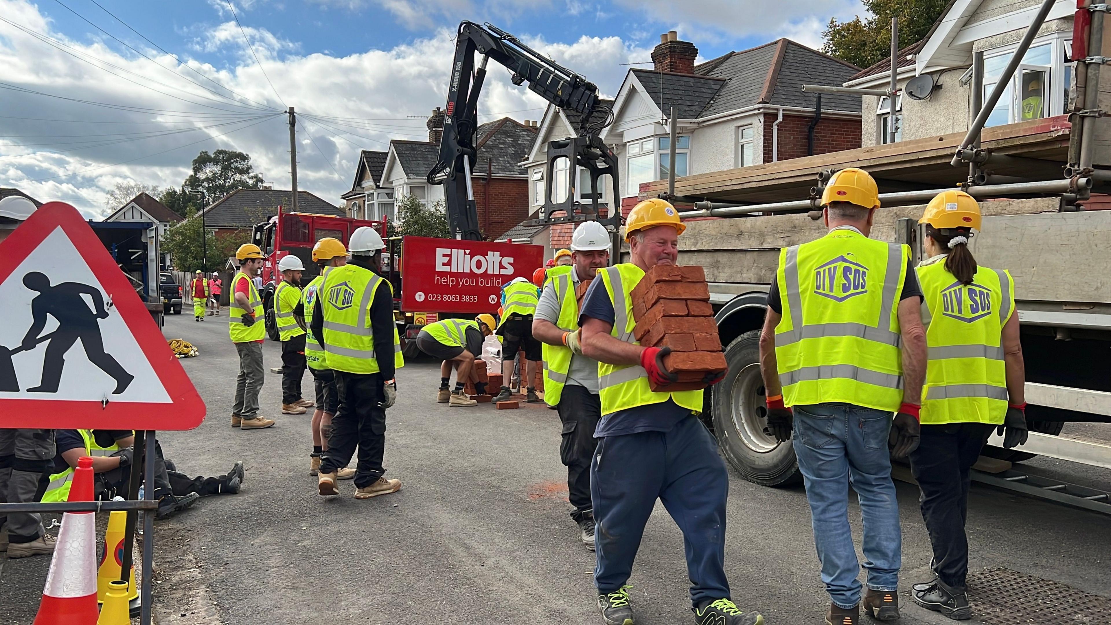 A street of men and women wearing florescent jackets and hard hats. There is a lorry unloading supplies and a man carrying a stack of red bricks. 