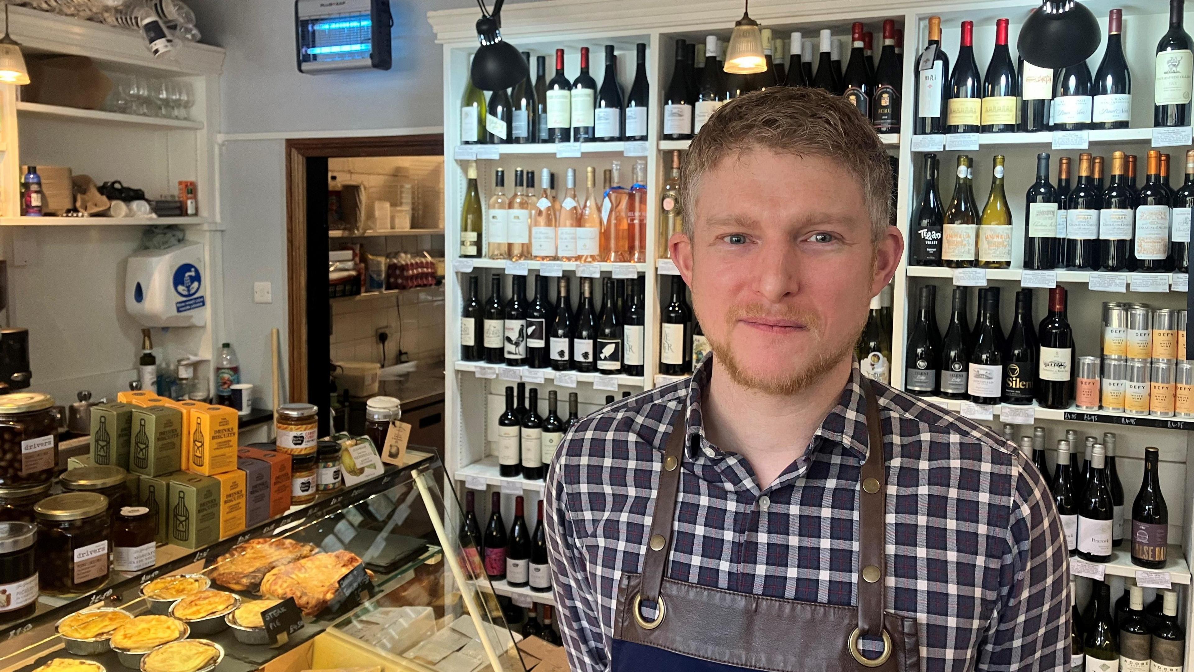 A man wearing a checked shirt and brown leather overall, smiles at the camera as he stands in a shop. There are pies in a counter to his left, with jams and other preserves on top of the counter, and wine bottles on a shelf behind him.