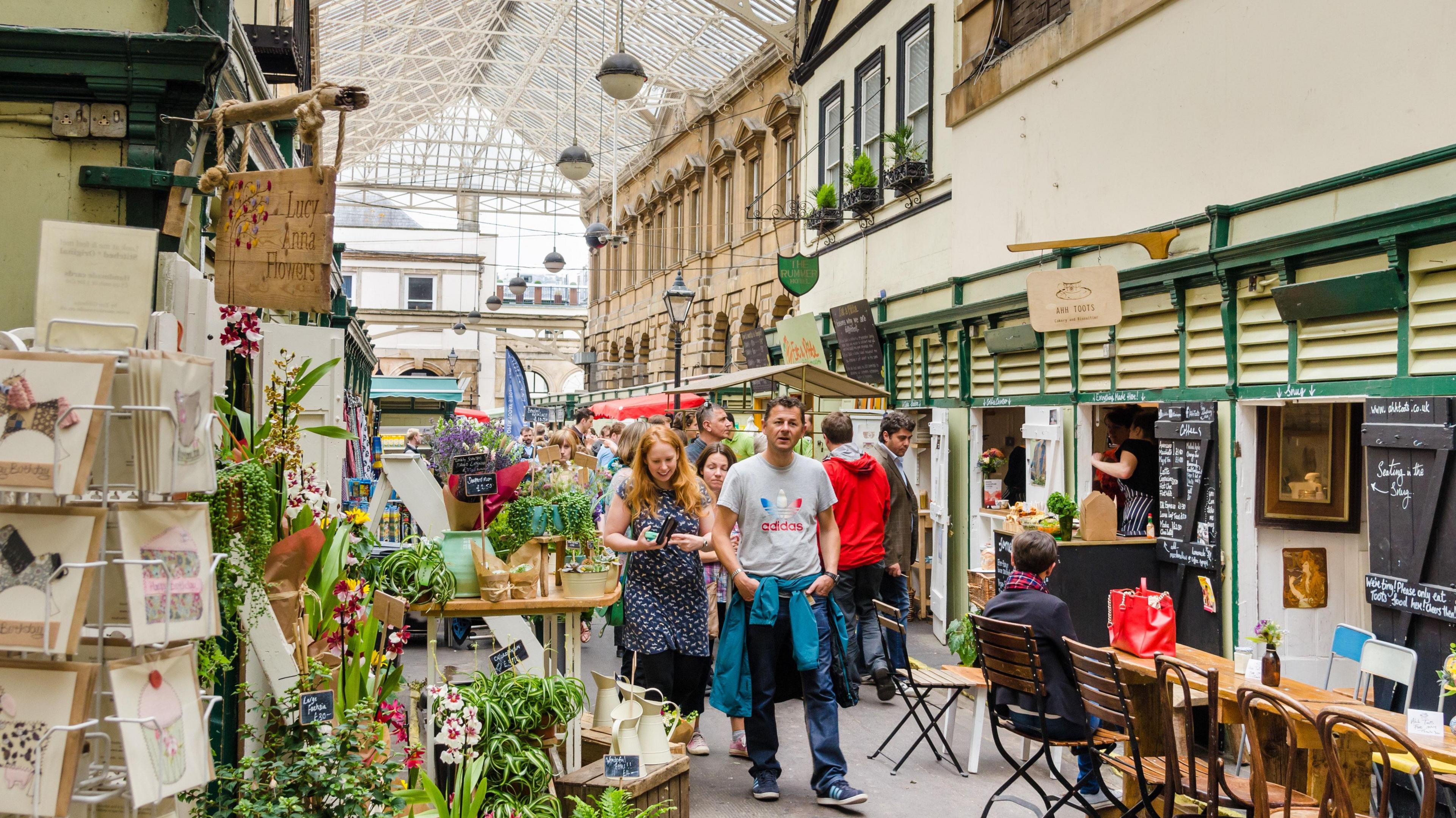 St Nicholas Market in Bristol. Customers can be seen walking through the indoor market, looking at vendors. Greeting cards and plants can be seen on the left hand side of the frame, while tables and a food stall can be seen on the right. 