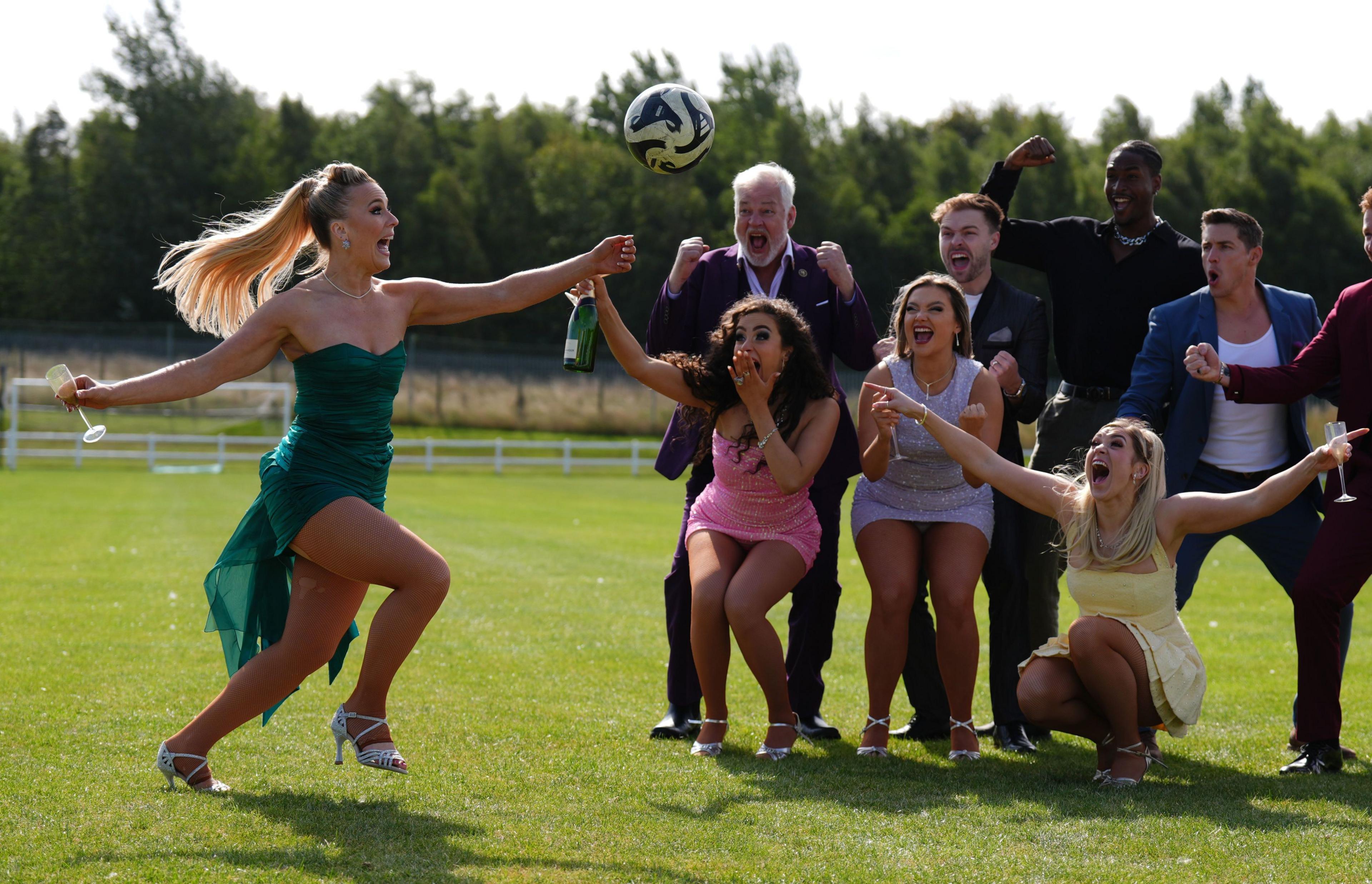 Ceili O'Connor, a blonde glamourously-dressed woman in a green dress and heels, kicks a football alongside fellow performers from Footballers Wives: The Musical - a group of men and women dressed in smart suits and dresses.
