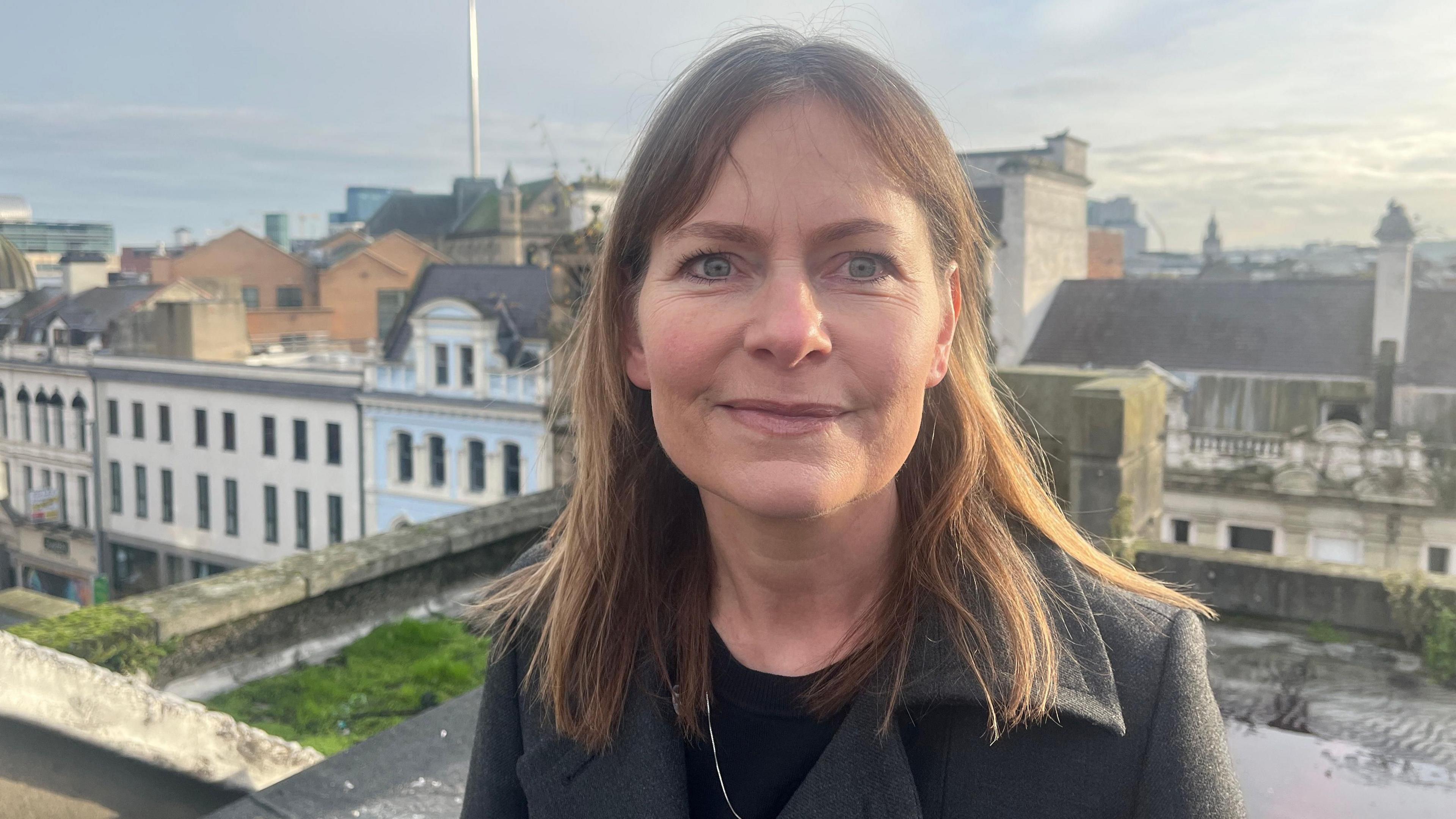 Wendy Langham - a woman with shoulder-length brown hair is standing on a rooftop and smiling at the camera. The picture shows her from the shoulders up. She is wearing a grey collared coat, a black top and a necklace.