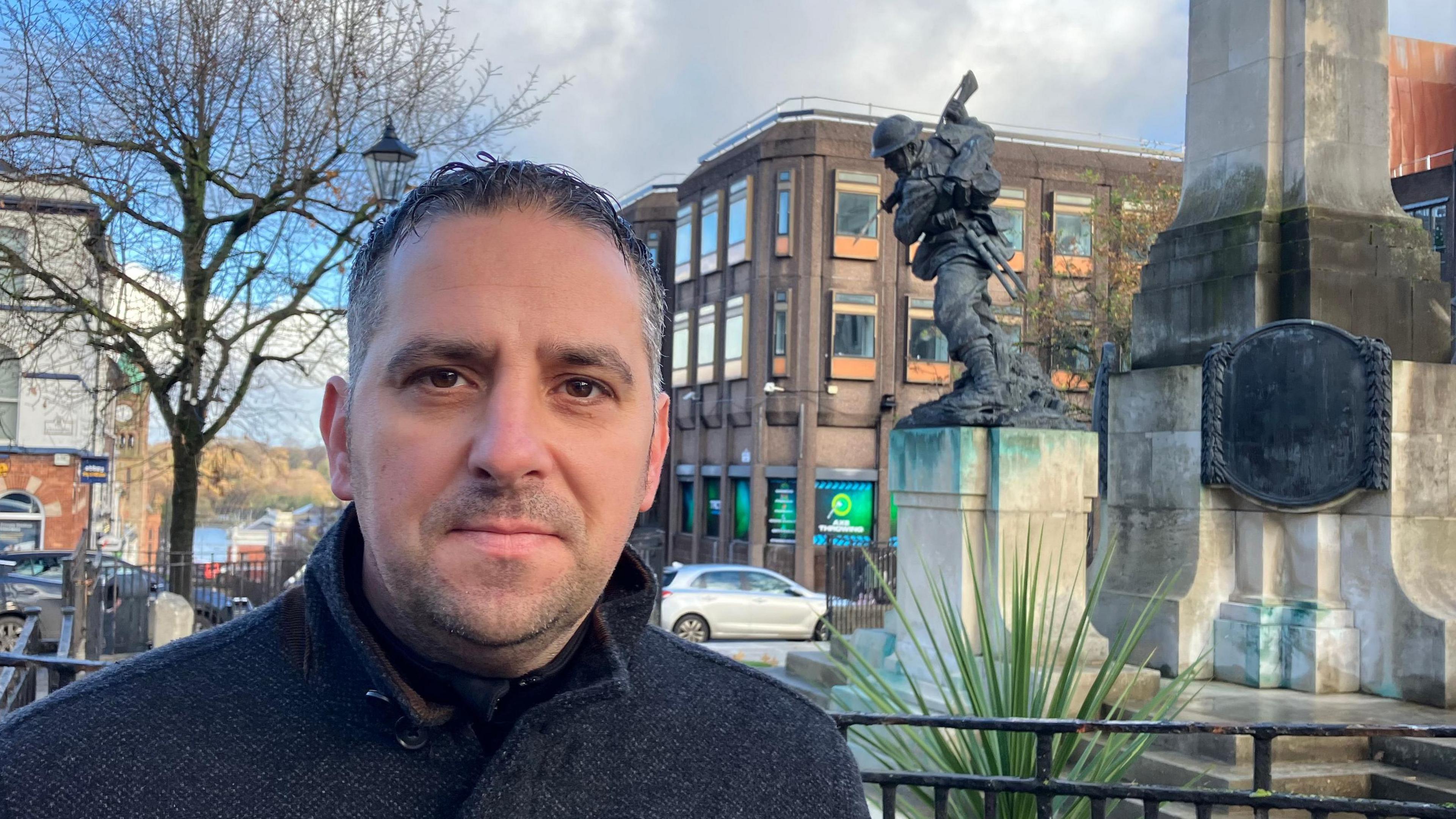 Christopher Jackson stands in front of the war memorial in Derry. He has short dark hair and is wearing a dark jacket, fully buttoned up