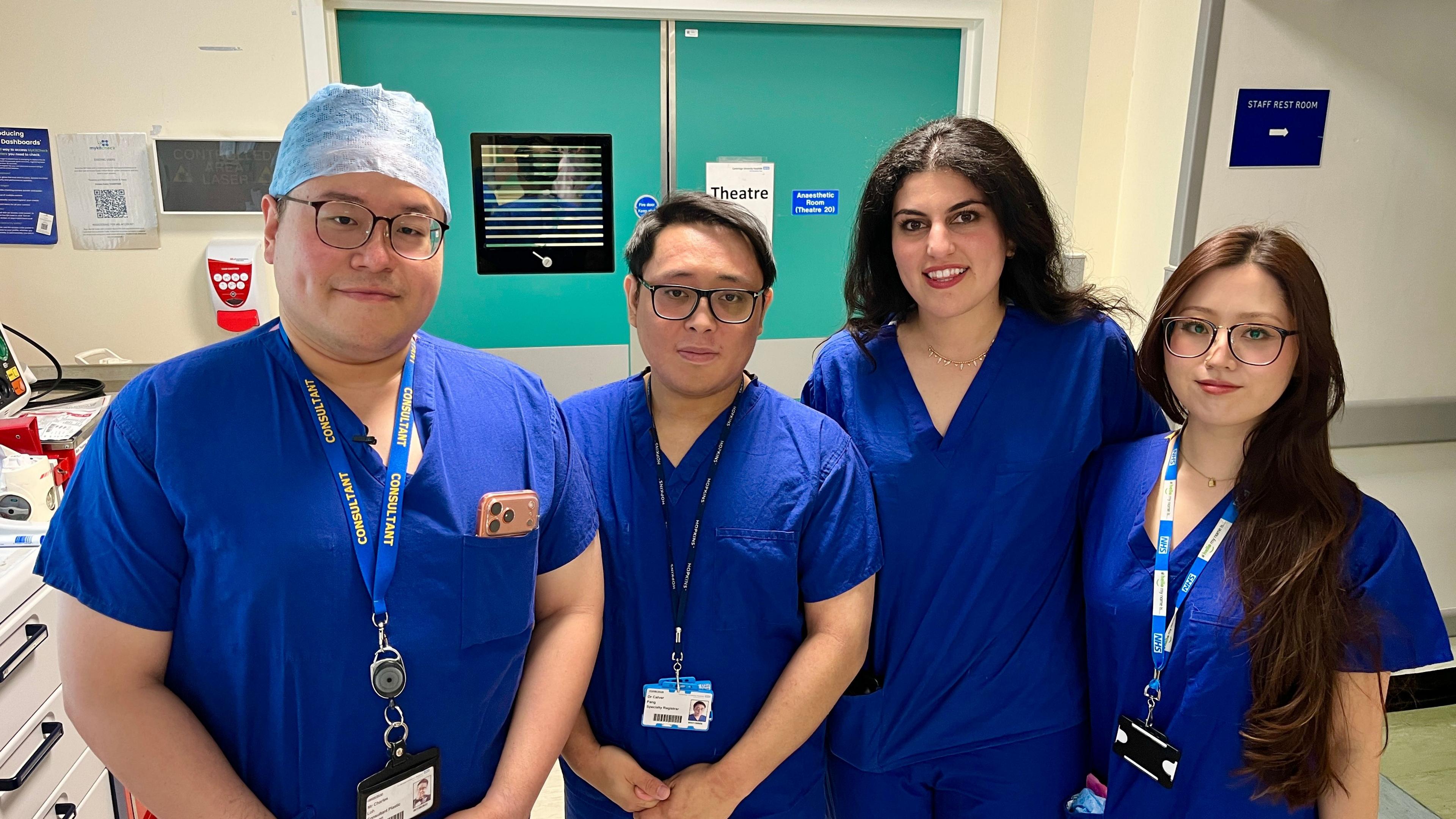 A team of clinicians in blue scrubs. Three have lanyards around their neck. They are standing in front of the doors to one of the theatres