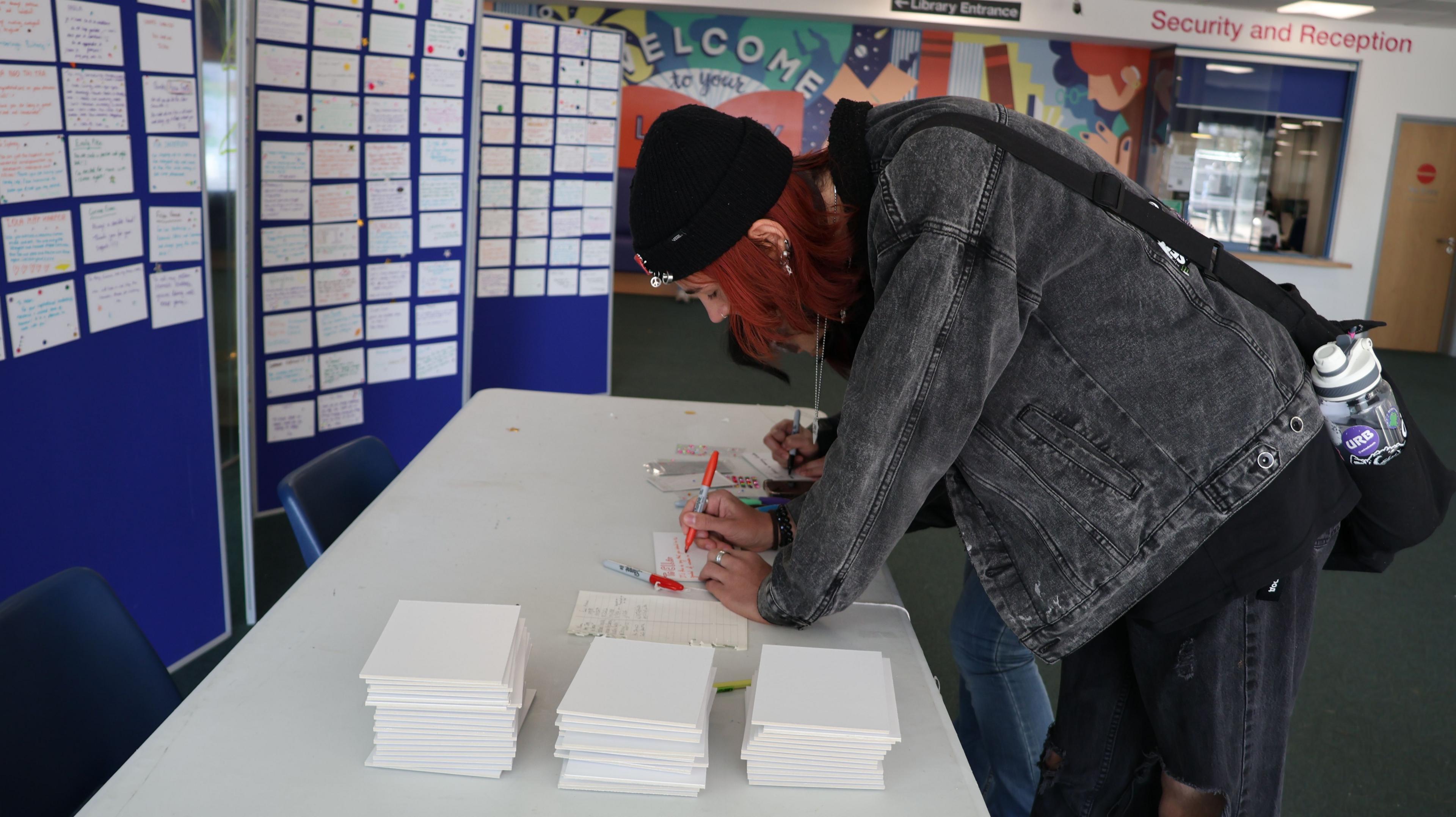 A woman with short red hair, wearing a black denim jacket, black beanie hat and jeans. She is leaning over a white table and writing on a small paper plaque with a red sharpie. In front of her are large blue display boards full of nomination plaques. 