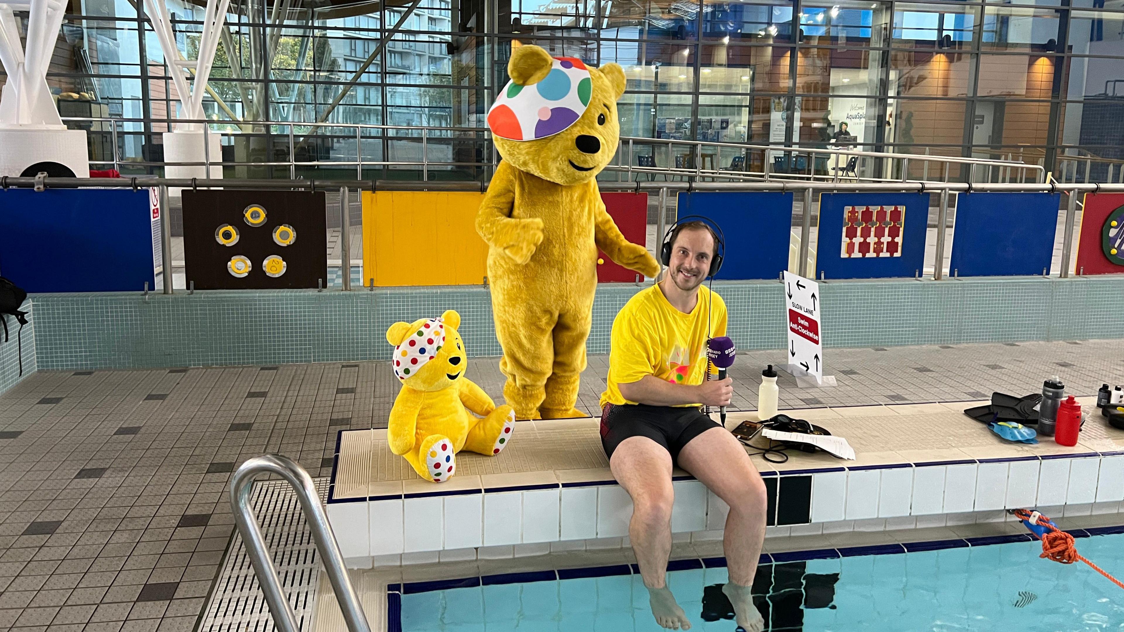 A reporter sits on the edge of the pool with his feet in the water while wearing a yellow top with Pudsey on it and black swimming trunks. He is holding a radio mic and has headphones on.  Pudsey dances behind him and there's a toy pudsey next to him.