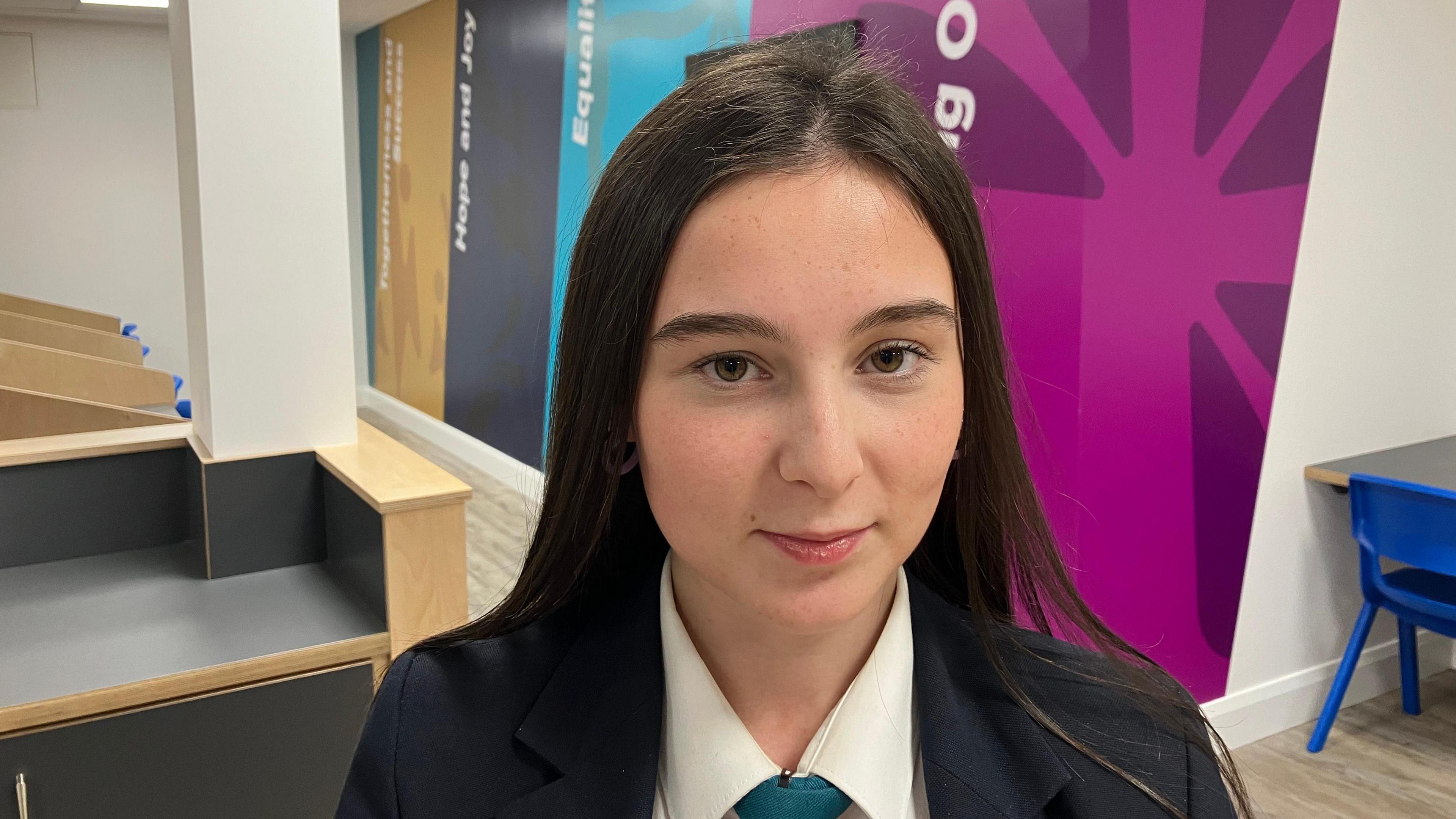 A school girl, pictured from the shoulders up. She is wearing a shirt and a blazer. She has long black hair. She is standing inside the school, with a white, purple and blue wall behind her. 