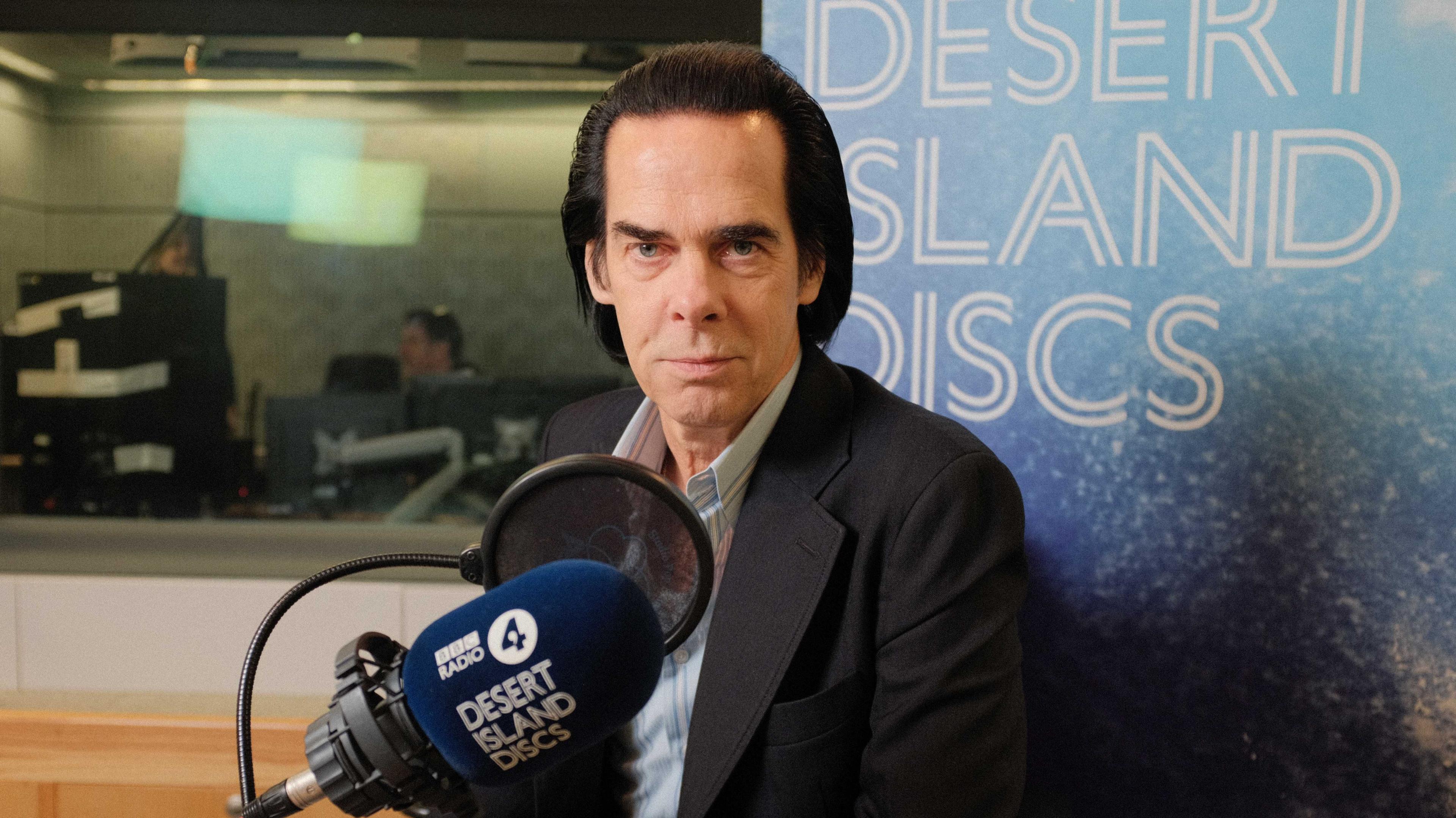 Nick Cave, with dark slicked back hair, wears a dark grey jacket and blue striped suit, in front of a sign reading Desert Island Discs, with a Radio 4 microphone in front of him 