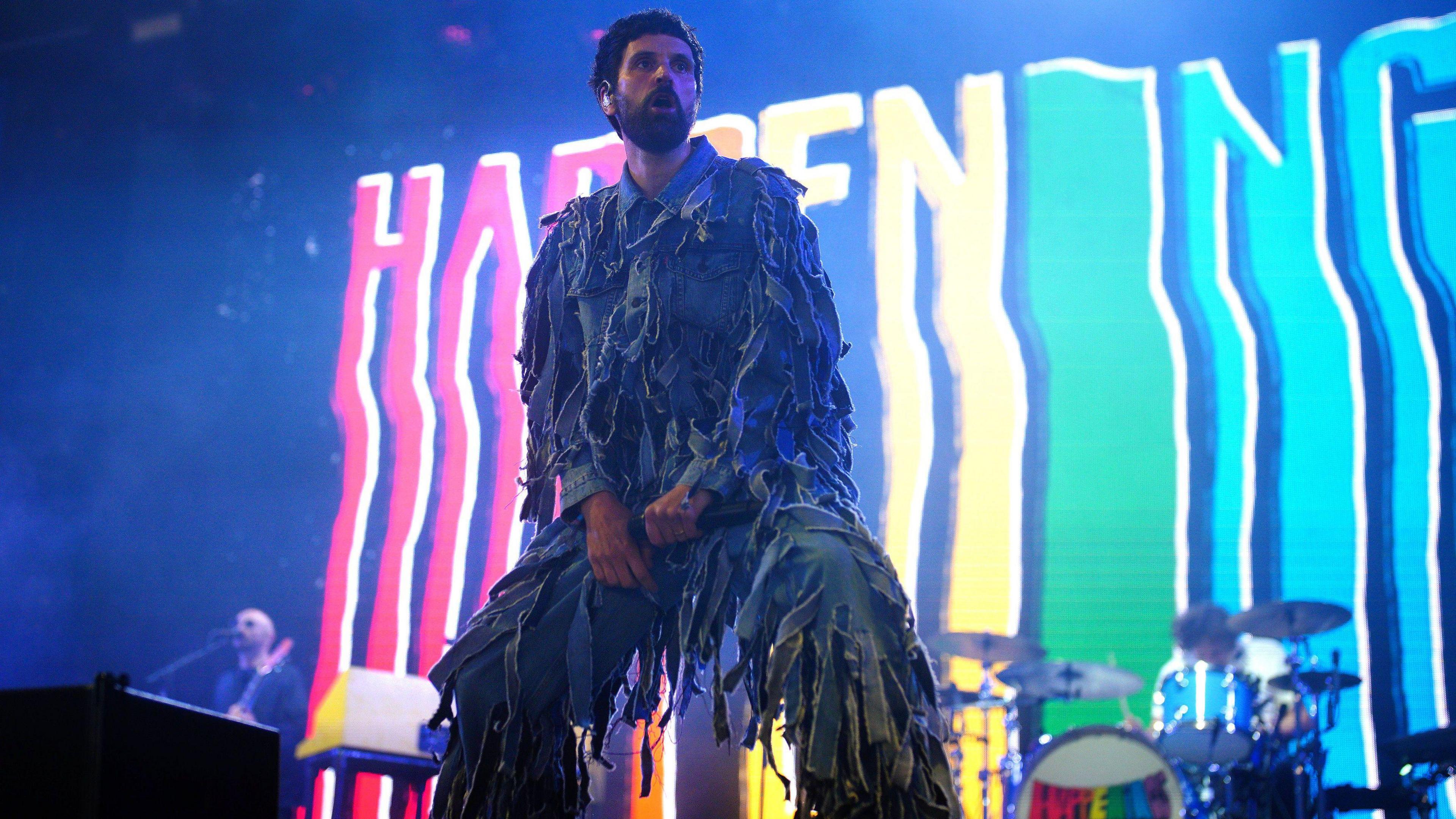 Serge Pizzorno from Kasabian. He has short dark hair and facial hair. He is pictured on stage, looking out at the crowd