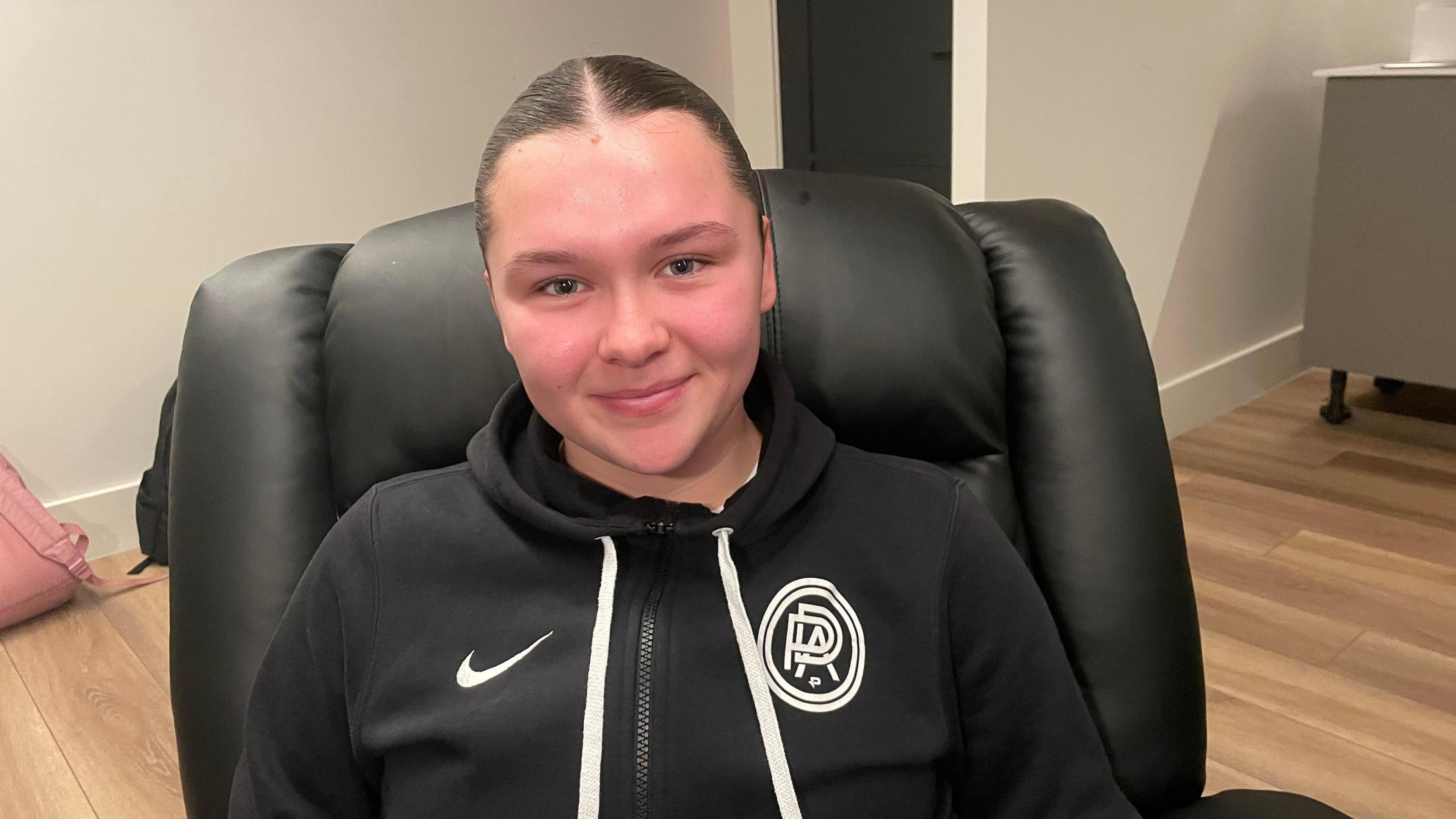 Picture of 17 year old college student Sophia, who has brown hair slicked back in a ponytail. She is sat on a black leather chair, smiling towards the camera and wearing a zip up black nike hoodie. 