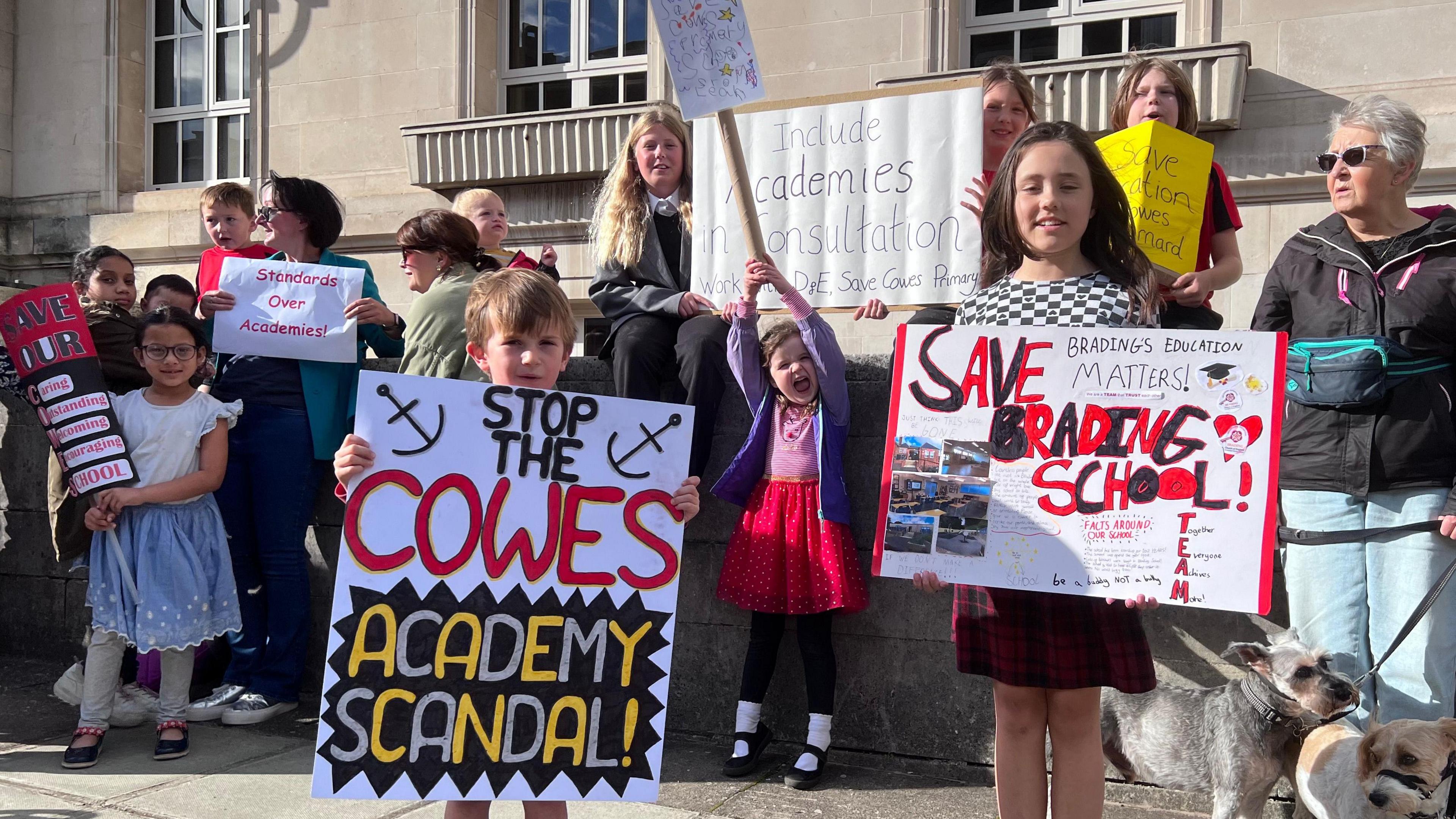 A group of children and adults standing outside County Hall in Newport. They hold signs with slogans including Stop the Cowes Academy Scandal, Save Brading school and Save our school