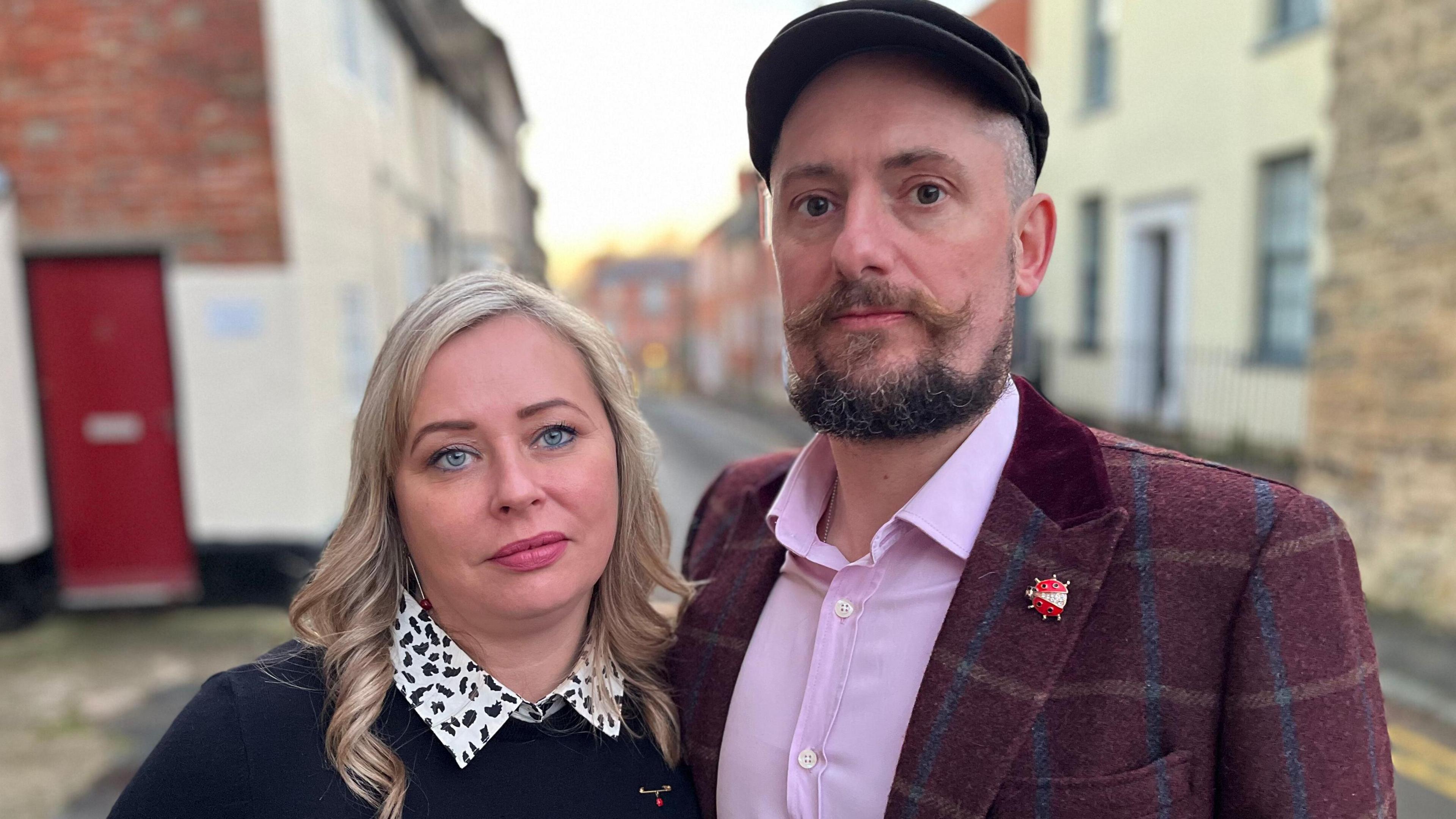 Couple Carly and Carl are pictured standing together in the street. There are terraced houses in the background. Carly has blonde hair and is wearing a black and white shirt and a dark jumper. Carl is wearing a cap and has a beard and moustache. He is wearing a lilac shirt with a maroon checked jacket. He has a ladybird brooch on the lapel.