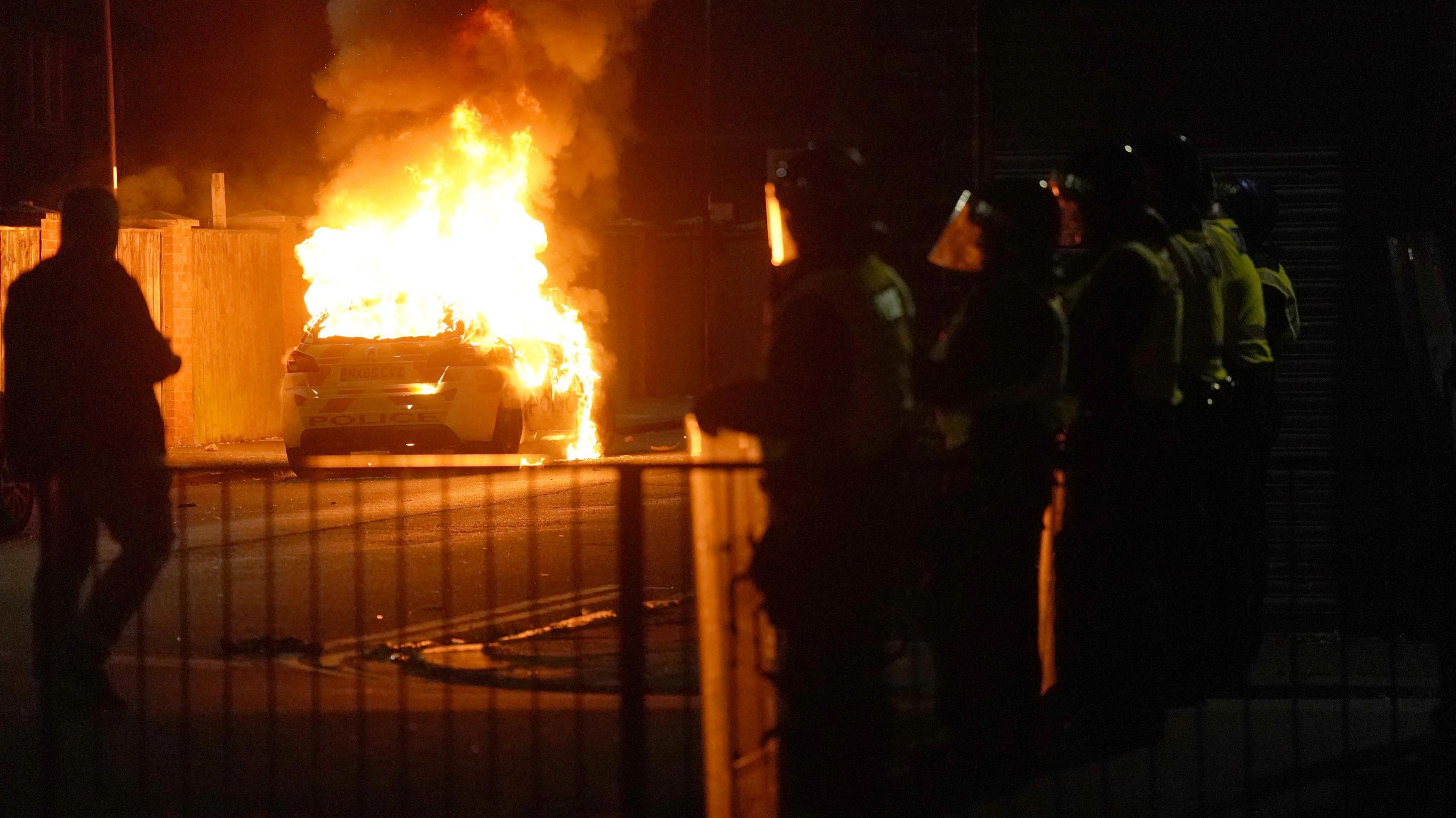 A police car burns with police officers lined on the street